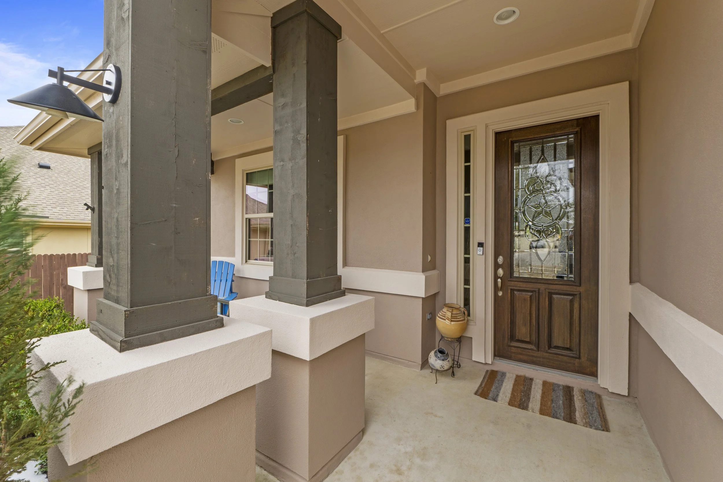 Close-up of a front porch with a decorative door, a small doormat, a tall vase, and a blue chair in the background, with columns supporting the porch roof.