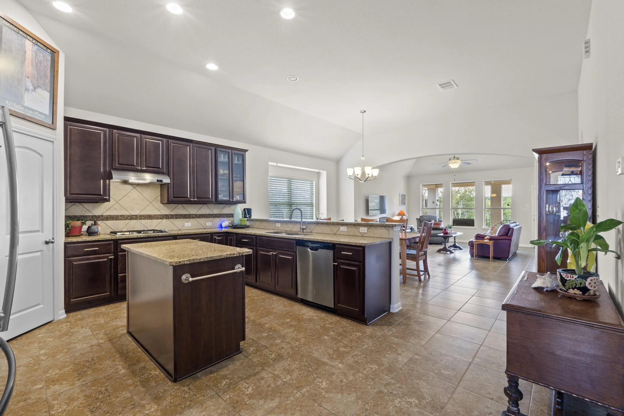 Open-concept kitchen and living room with dark wood cabinets, beige tile flooring, and a kitchen island in the foreground, flowing into a dining area with a chandelier and a living area with chairs, a sofa, a TV, and large windows bringing in natural