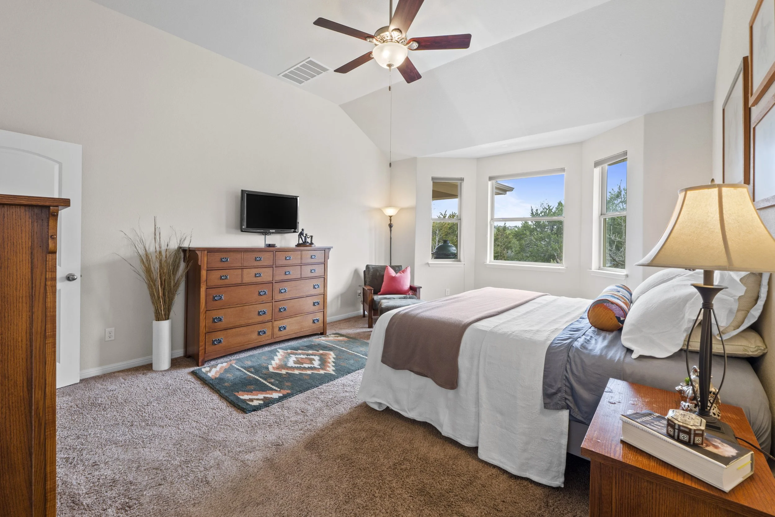 A bedroom with a bed, window view of trees, wooden furniture including a dresser with a TV, a bedside table, a chair with a pink cushion, and a ceiling fan.