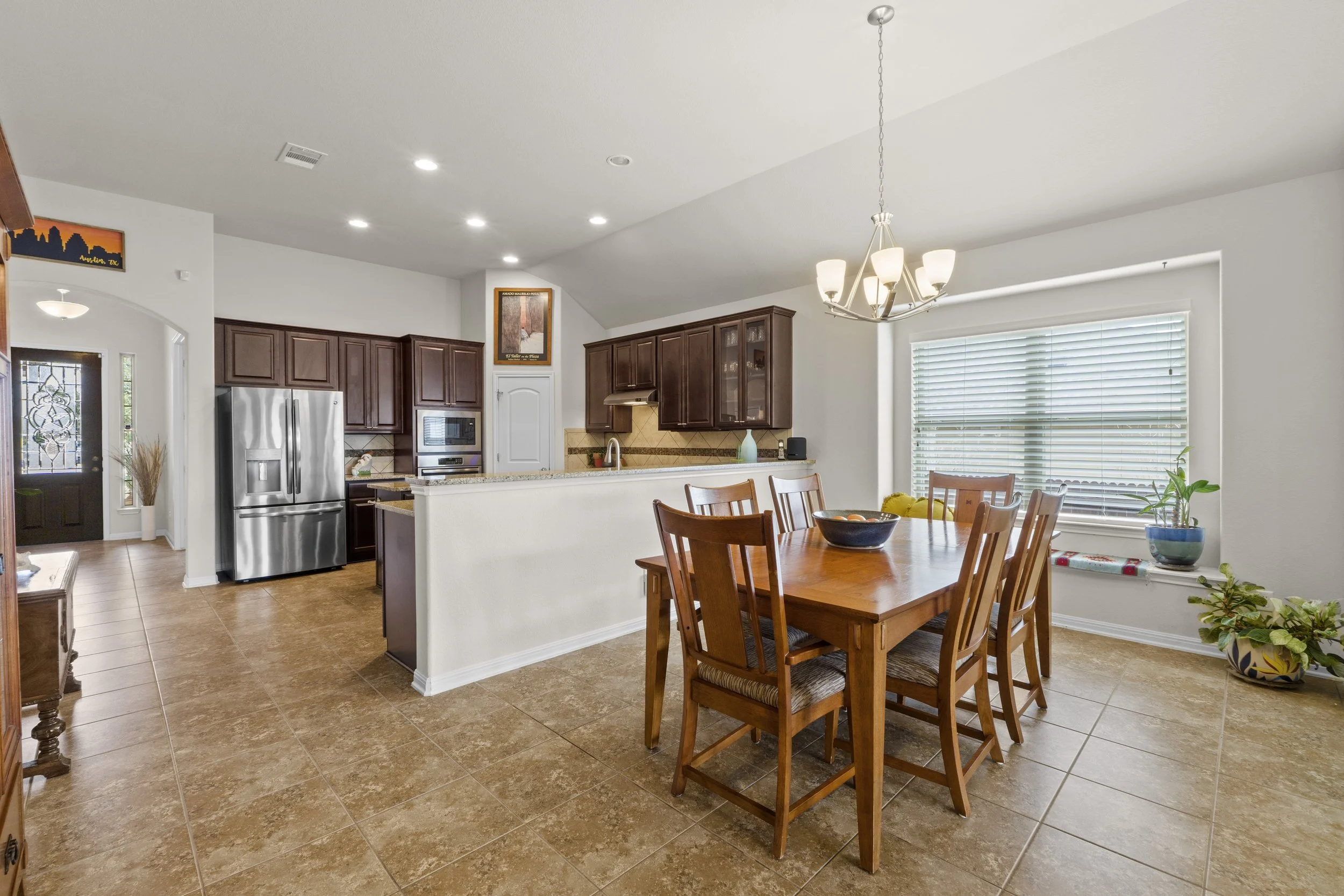 A dining area with a wooden table and six chairs, a window with blinds, a chandelier above, and a view into a kitchen with brown cabinets, stainless steel refrigerator, and microwave.