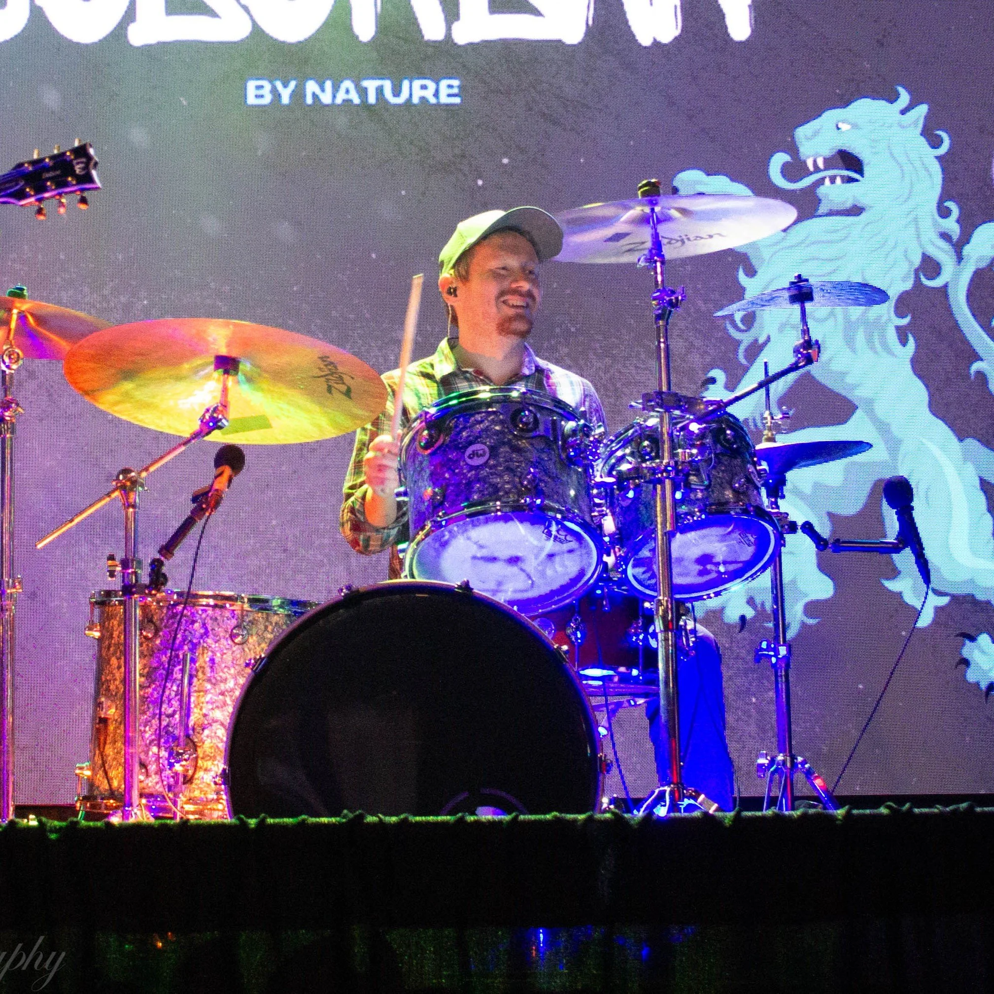 Jason of Suburban by Nature playing drums on a stage with colorful lighting and a large digital screen in the background displaying a blue creature and the text "BY NATURE".