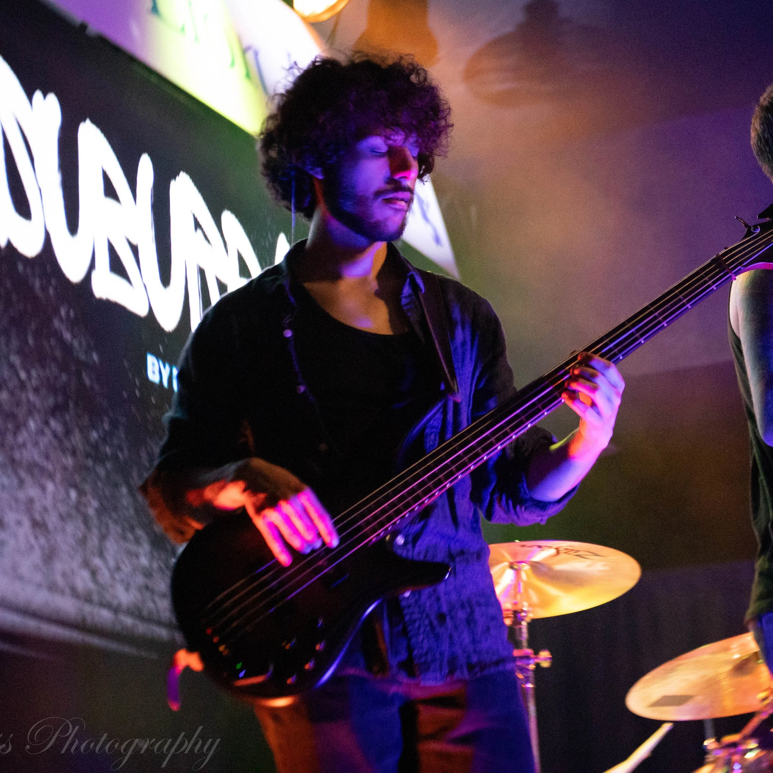 Felix of of Suburban by Nature playing bass at an event with colorful lighting and a sign in the background.