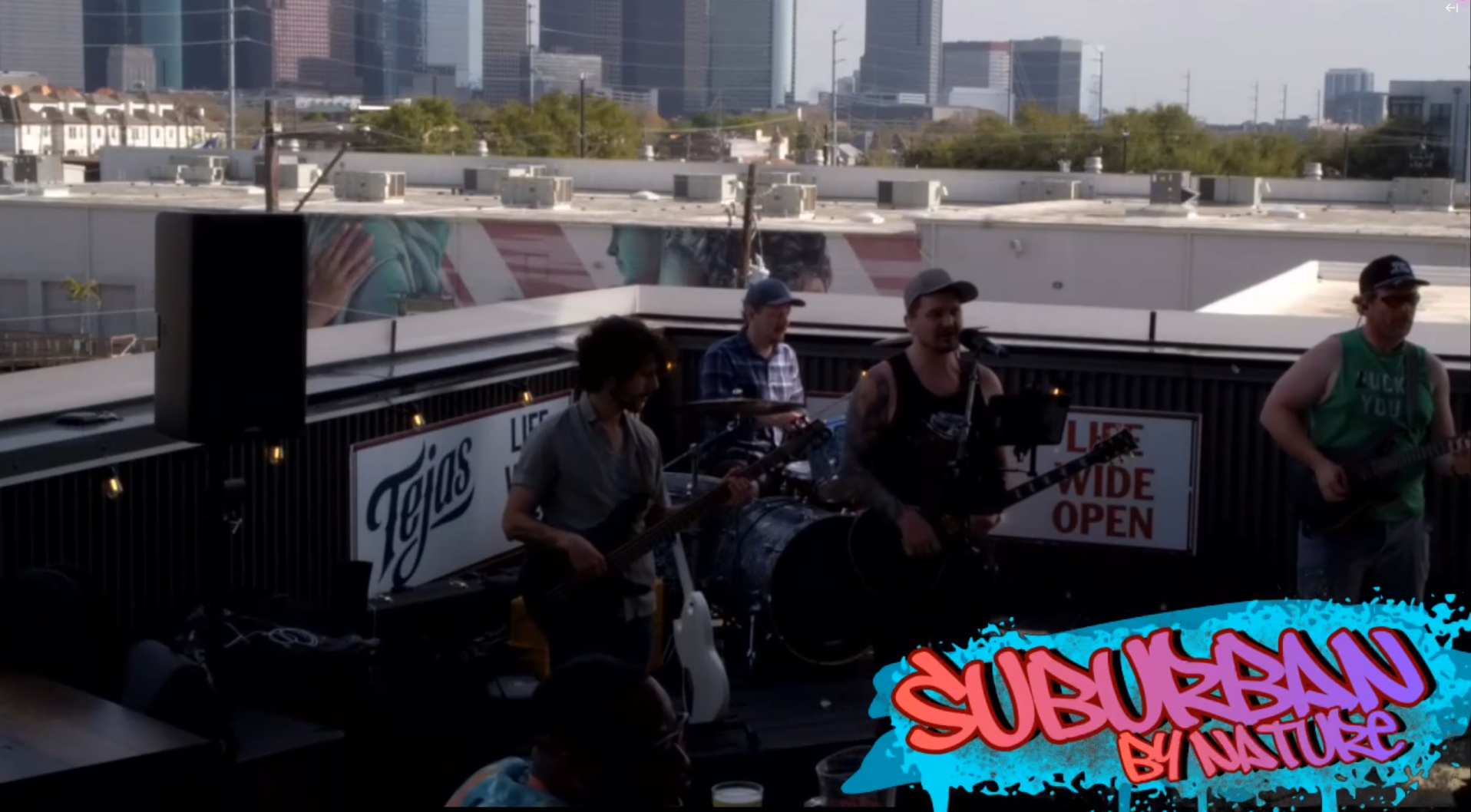 Suburban by Nature performing on an outdoor rooftop stage with city skyscrapers in the background, featuring Casey Bowen and 3 musicians playing guitars and drums, and a colorful banner that reads 'SUBURBAN by NATURE' on the stage.
