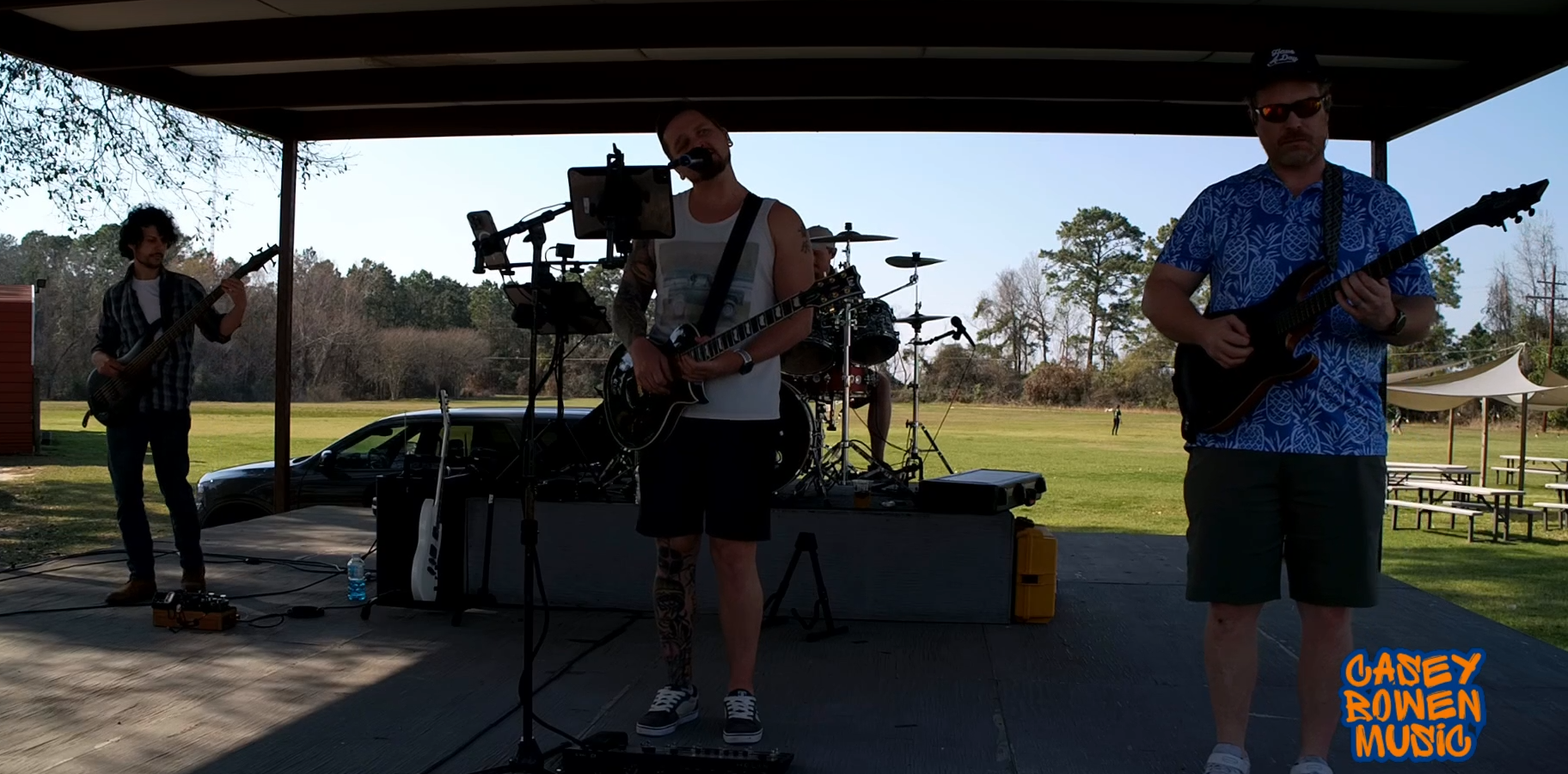 A band performing outdoors under a canopy, with a bassist on the left, a drummer in the middle, and a guitarist on the right. The background shows a grassy field, trees, and picnic tables. The image includes a logo in the bottom right corner reading 'Casey Rowan Music'.