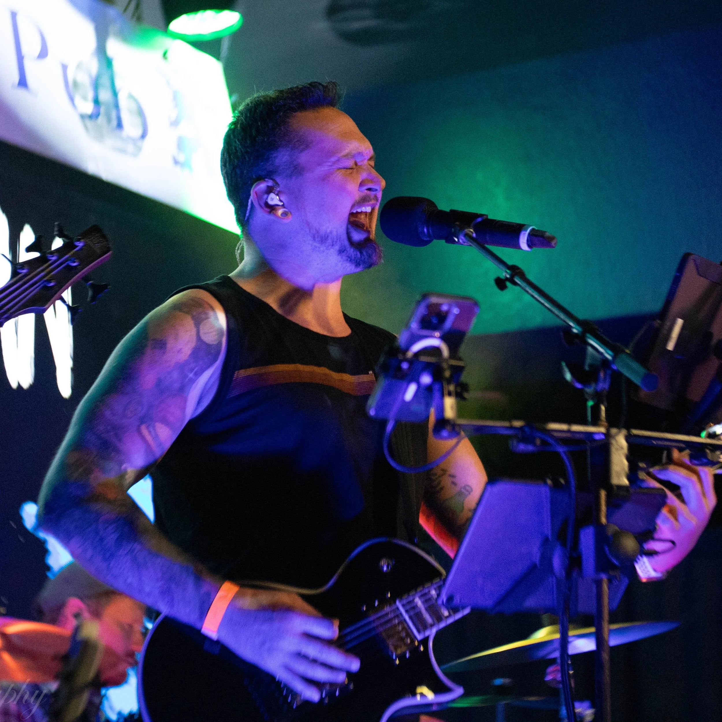 Casey Bowen of Suburban by Nature singing into a microphone at a live music performance, illuminated by colorful stage lights.