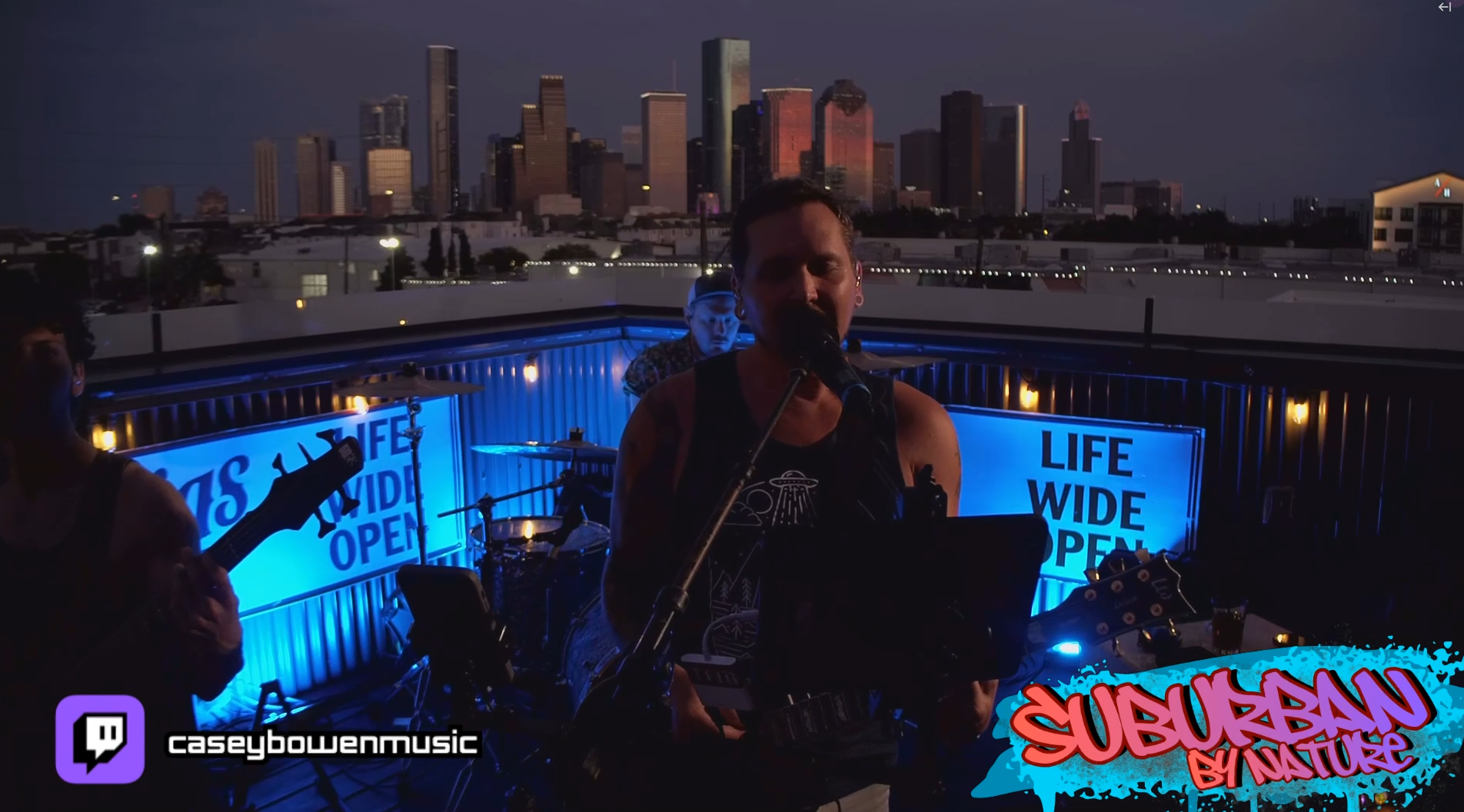 A rooftop music performance at night with a city skyline in the background, featuring Casey Bowen and Suburban by Nature, illuminated by blue and yellow lights, with a graphic saying "SUBURBAN BY NATURE."