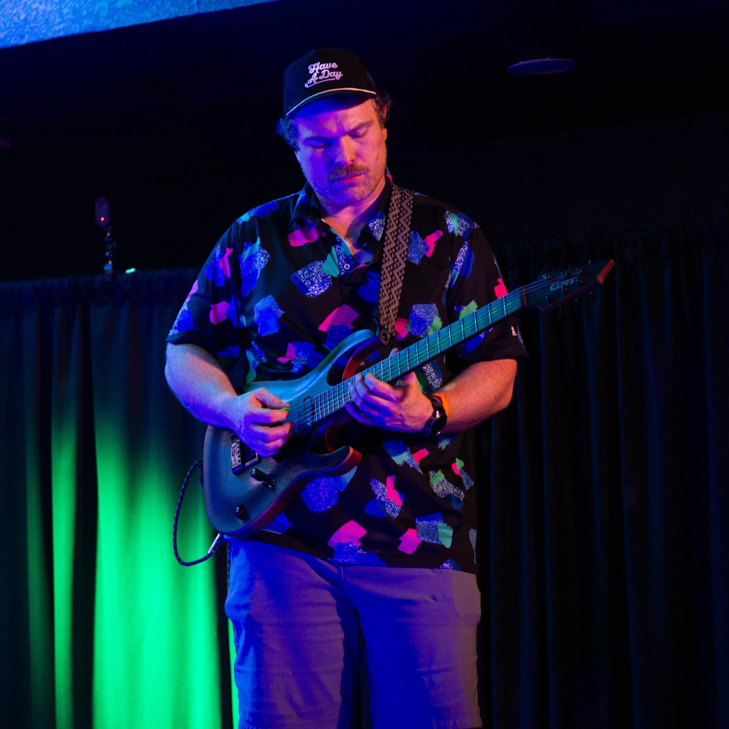 Tommy of Suburban by Nature playing an electric guitar on stage under colored lights, wearing a black cap with white text that says 'Have A Day' and a patterned shirt.