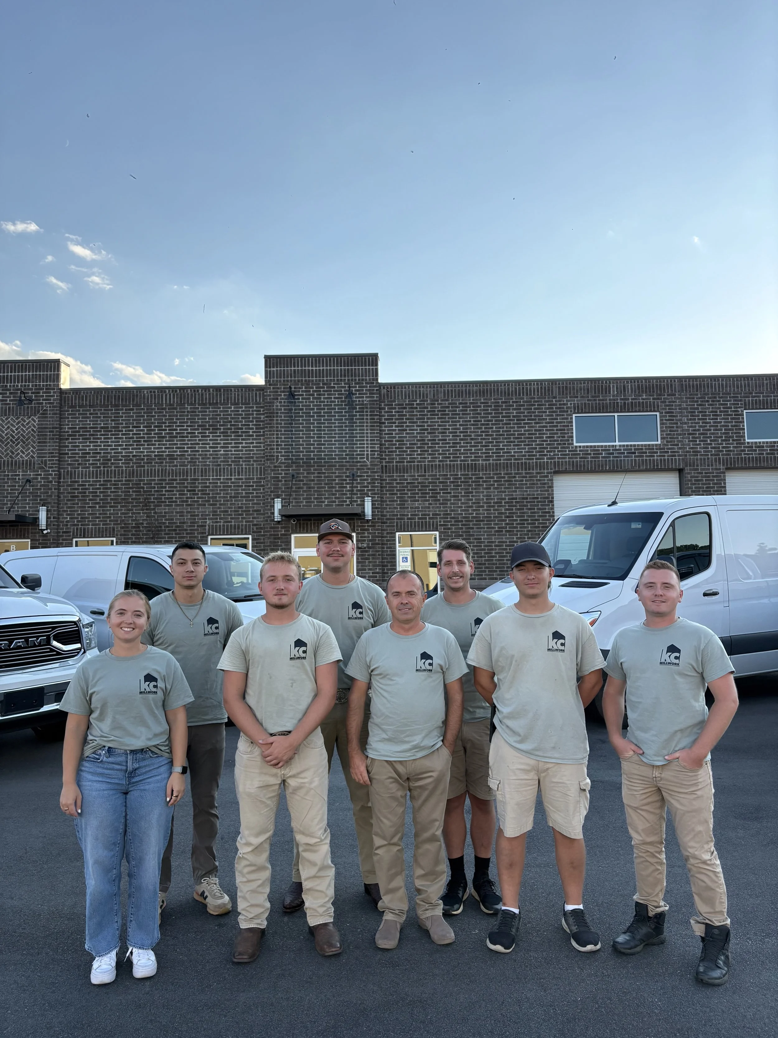 Group of eight people standing outside in front of vans and a brick building, wearing matching gray t-shirts with the KC company logo, under a blue sky.