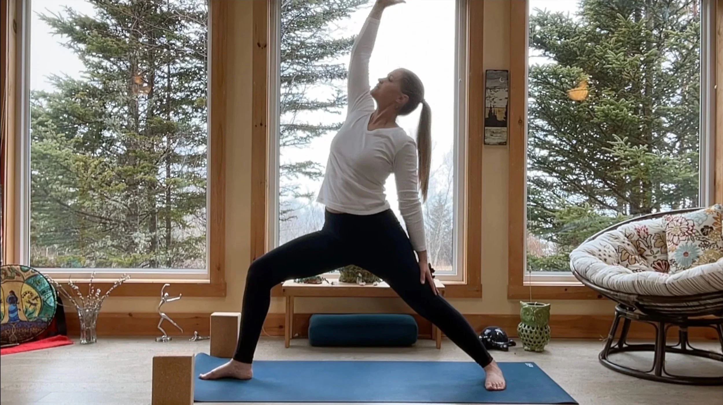 A woman practicing yoga indoors on a blue mat, performing a Warrior pose with an open stance, one arm reaching upward, and her gaze directed upward, in front of large windows showing evergreen trees outside.