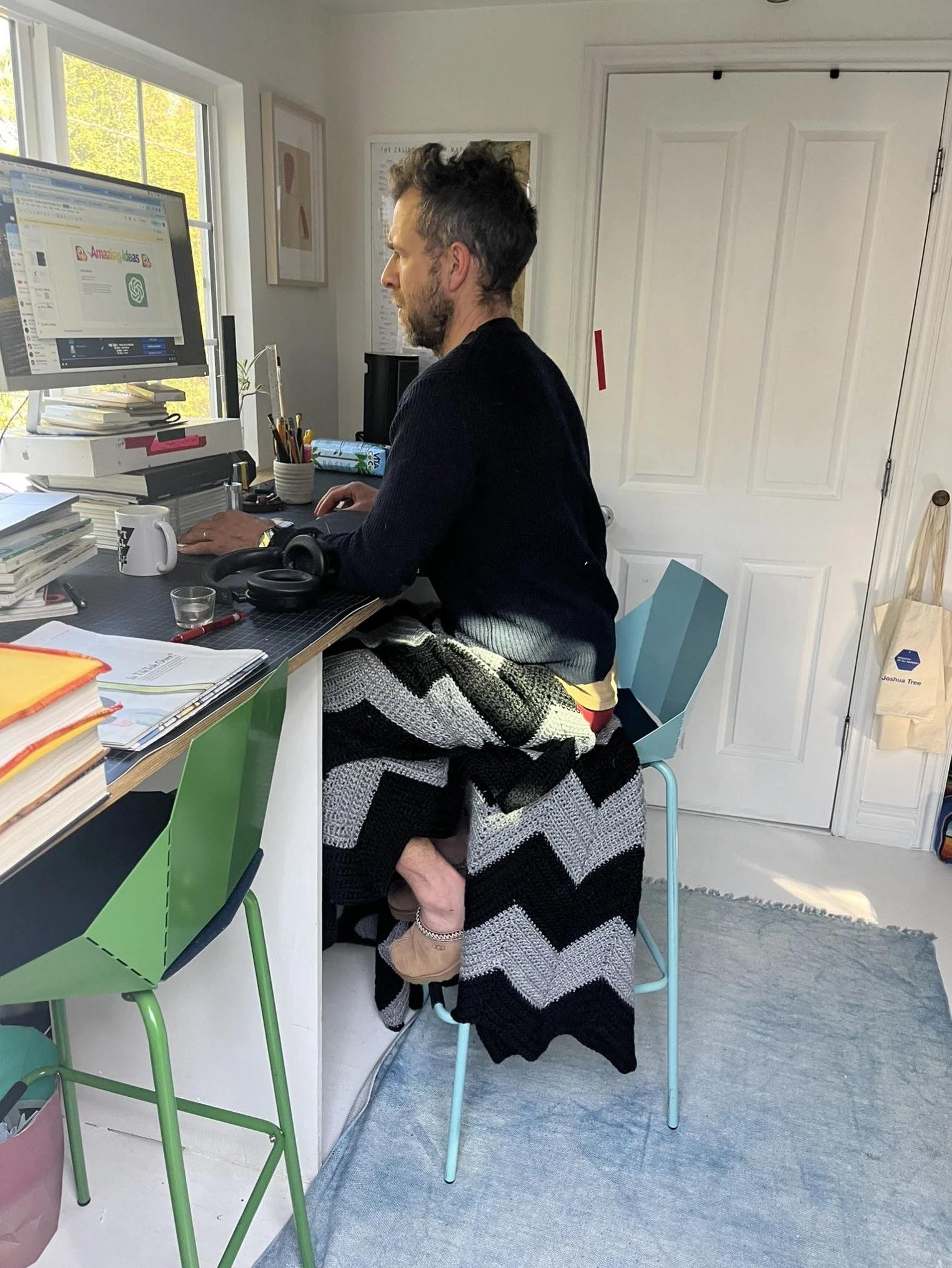 A man and a woman are in a bright home office or studio. The man is working on a computer at a cluttered desk, and the woman is leaning on the man's leg, wearing a black and white striped knit cardigan. The desk is filled with books and office supplies. The room has a window with sunlight, framed art on the wall, and a white door in the background.