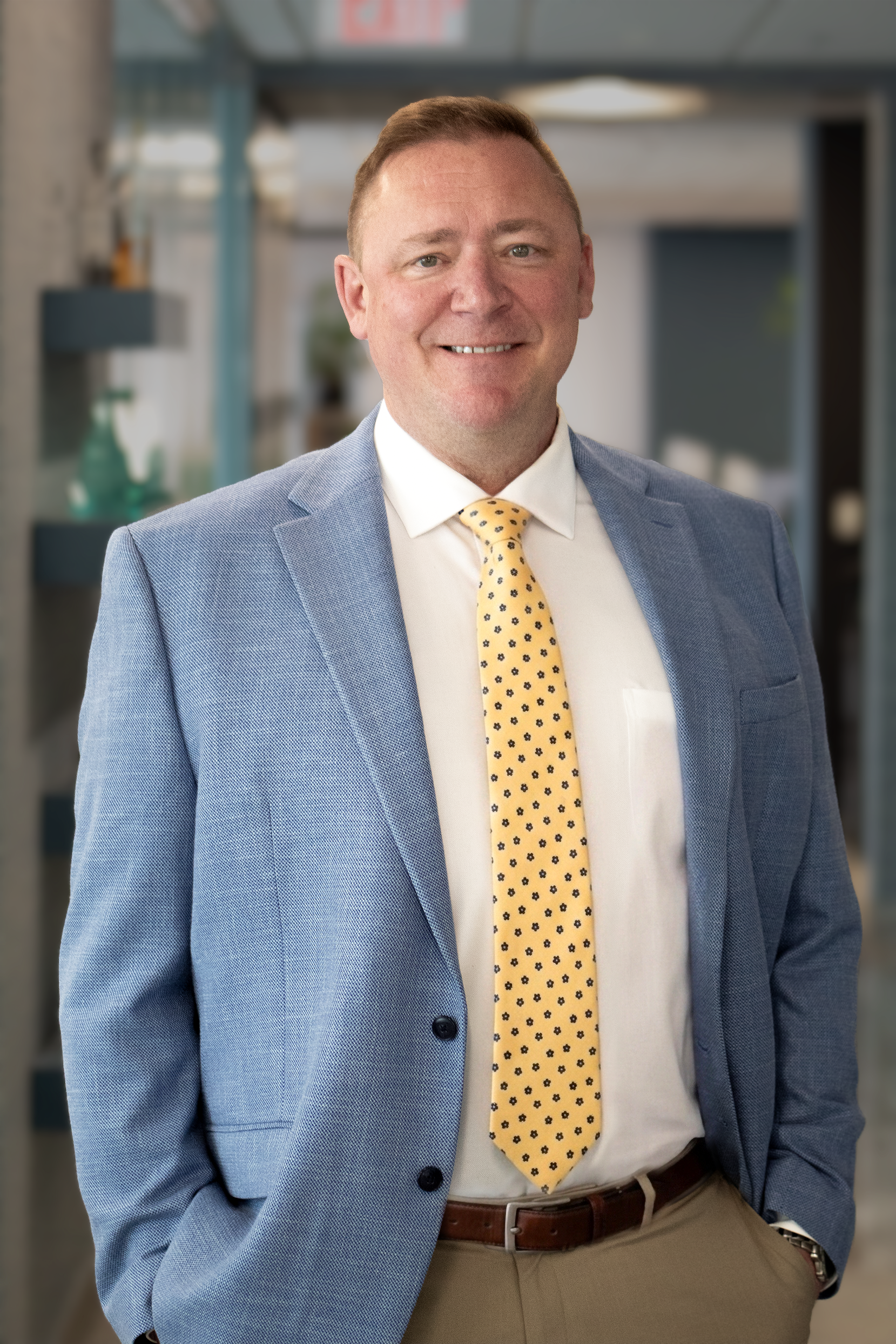 A man in a blue suit jacket, white shirt, and yellow tie with small dark patterns, standing with hands in pockets against an office background, smiling.