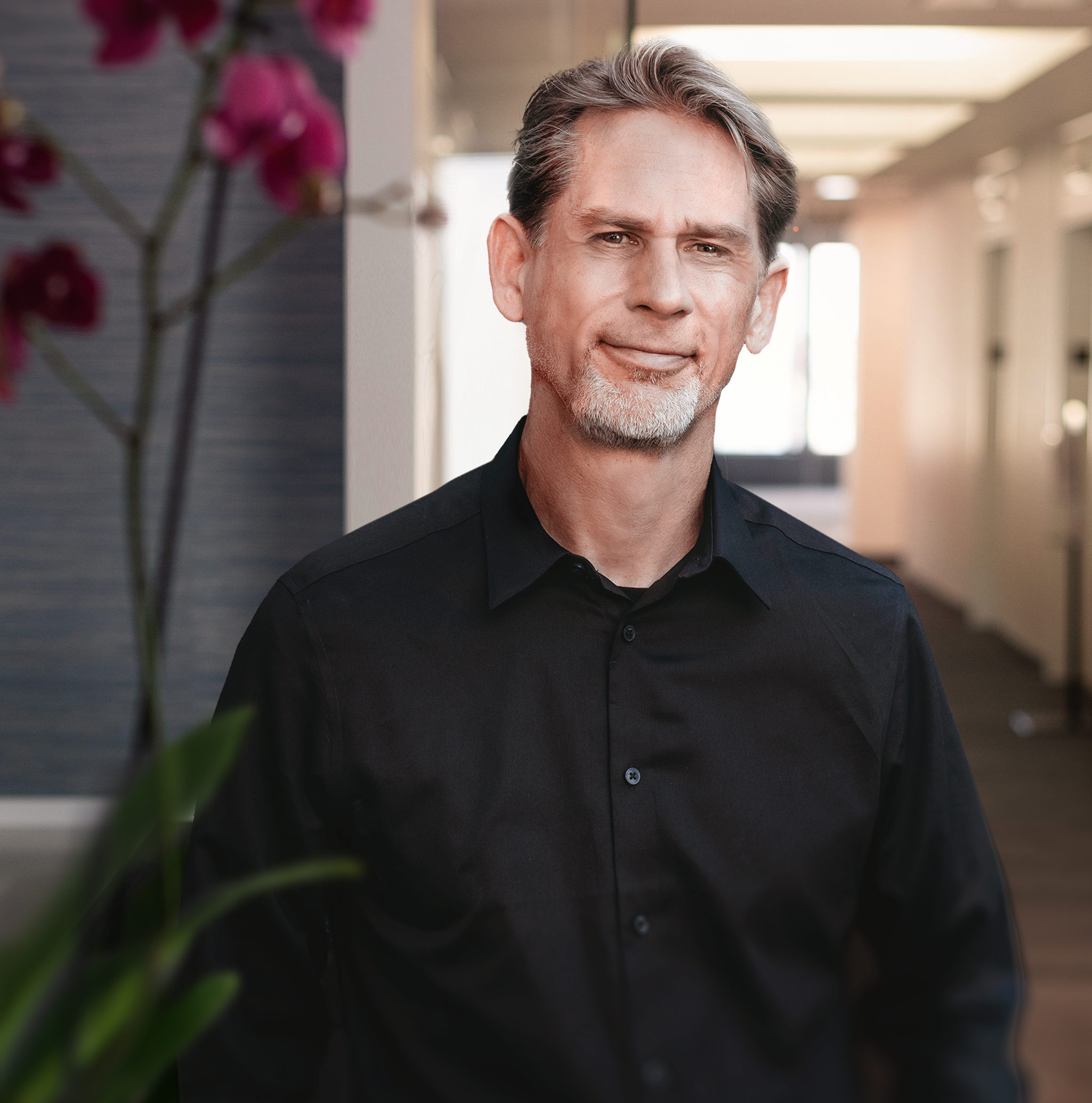 Close-up portrait of a middle-aged man with gray hair and beard, wearing a black shirt, standing indoors with natural light coming through the windows, blurred background, and a purple flower in the foreground.