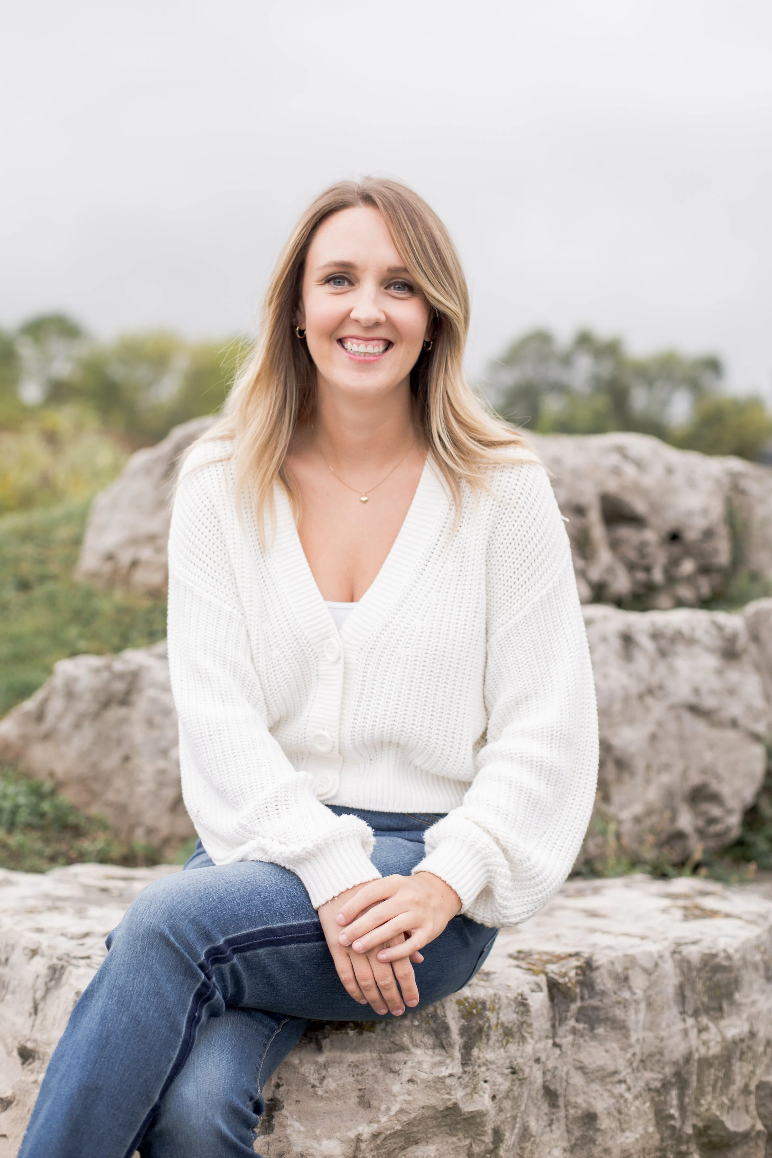 A smiling woman in a white sweater and blue jeans seated on rocks outdoors with greenery in the background.