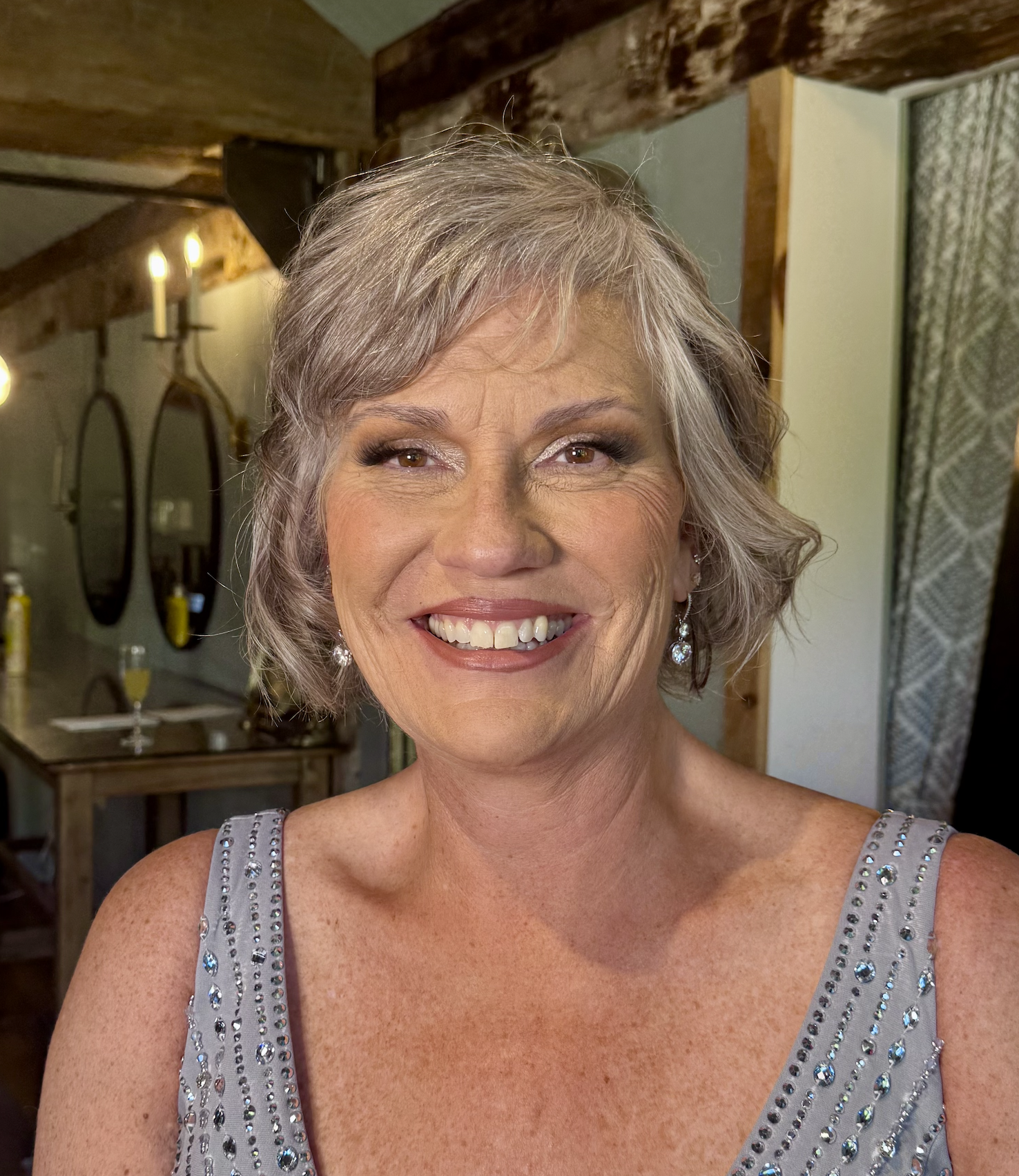 A smiling woman with short gray hair, wearing earrings and a silver dress with rhinestone embellishments, in an indoor setting with wooden beams and candles in the background.