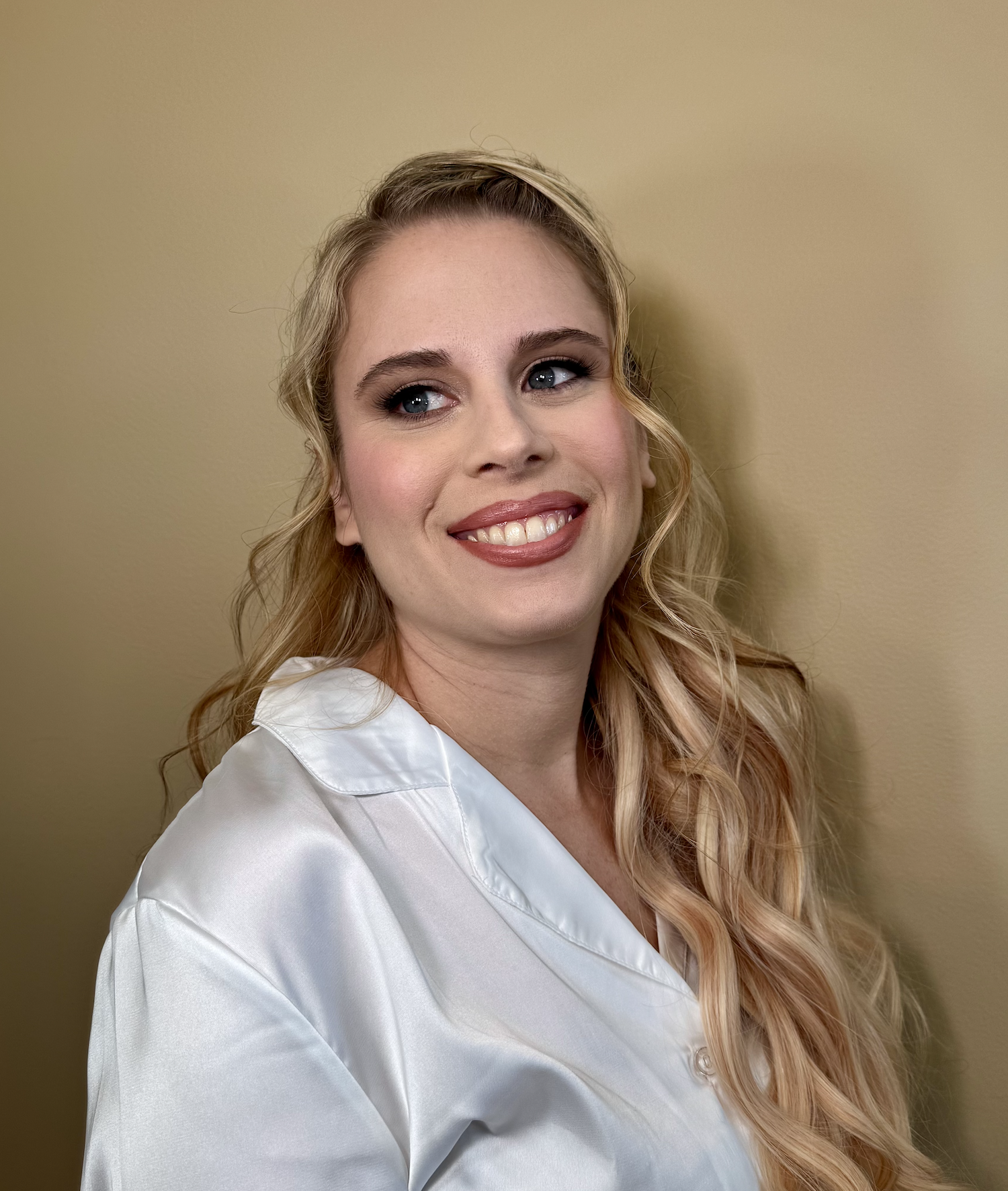 A woman with blonde wavy hair and blue eyes smiling at the camera, wearing a white satin blouse, standing against a beige background.