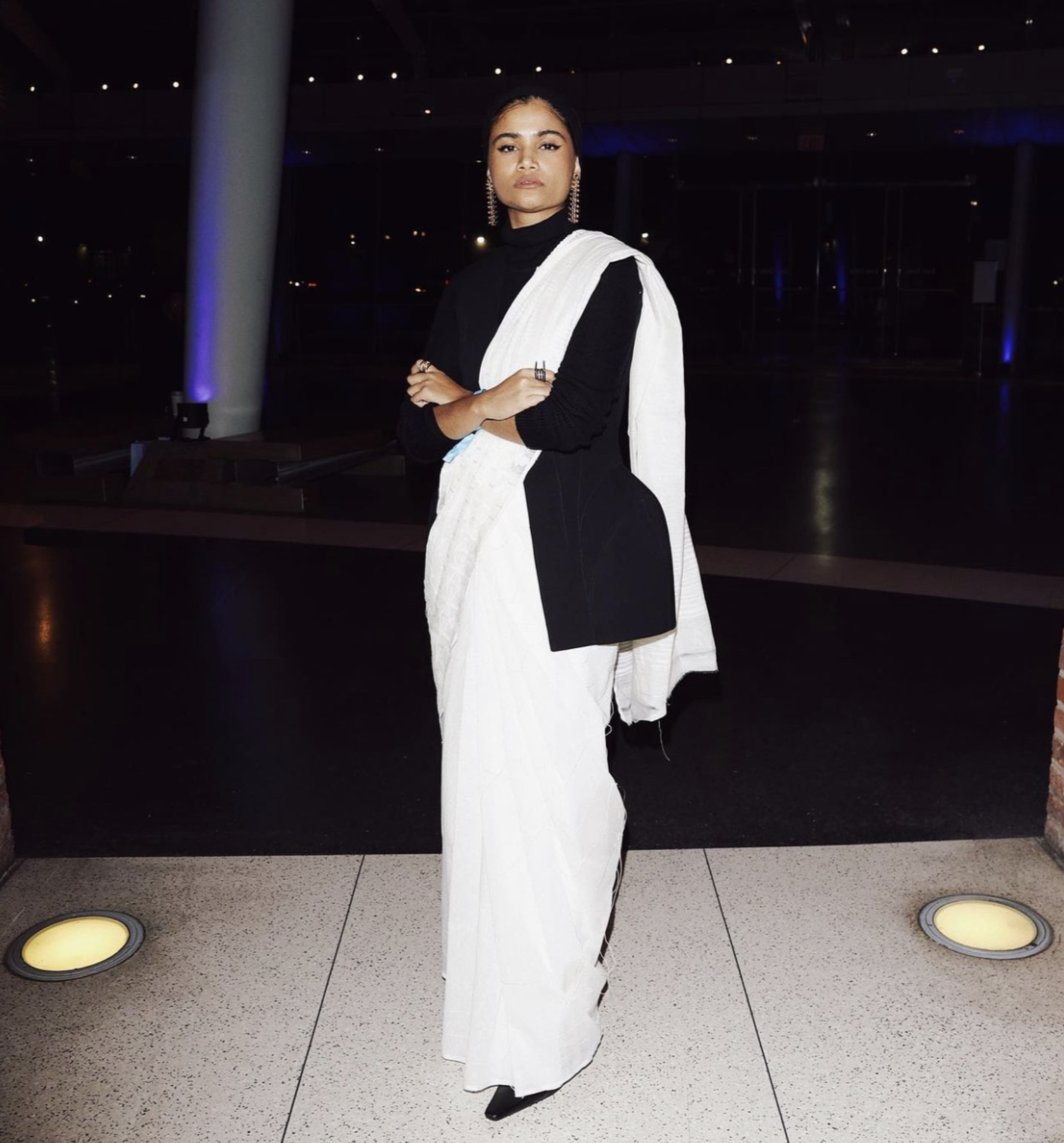 A woman standing indoors at night, wearing a black top, white sari, black shoes, and earrings, posing with her arms crossed. The background is dark with some blue and purple lighting.