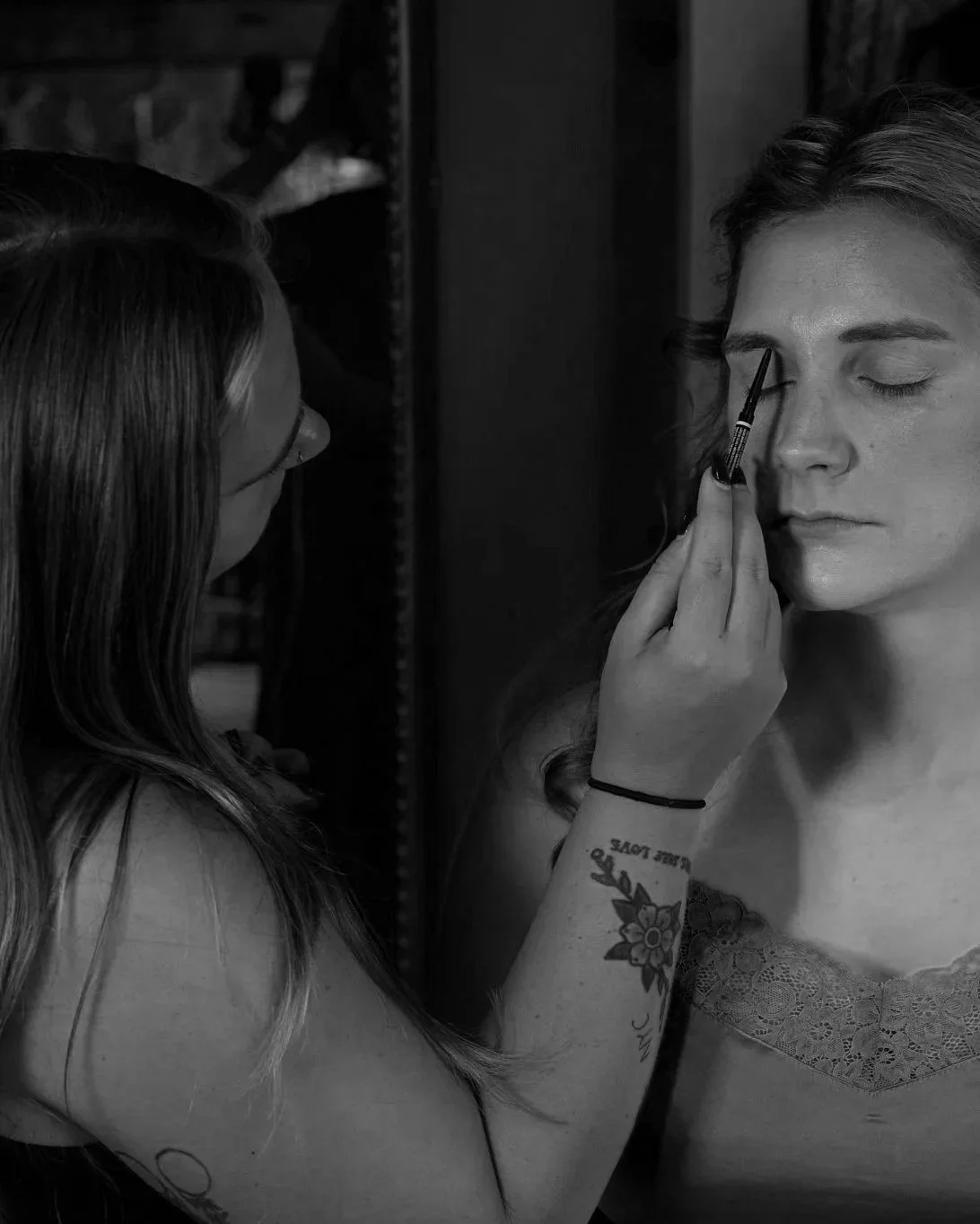 A woman applying makeup to another woman’s eyebrows, with one woman holding a makeup brush, in a monochrome setting.
