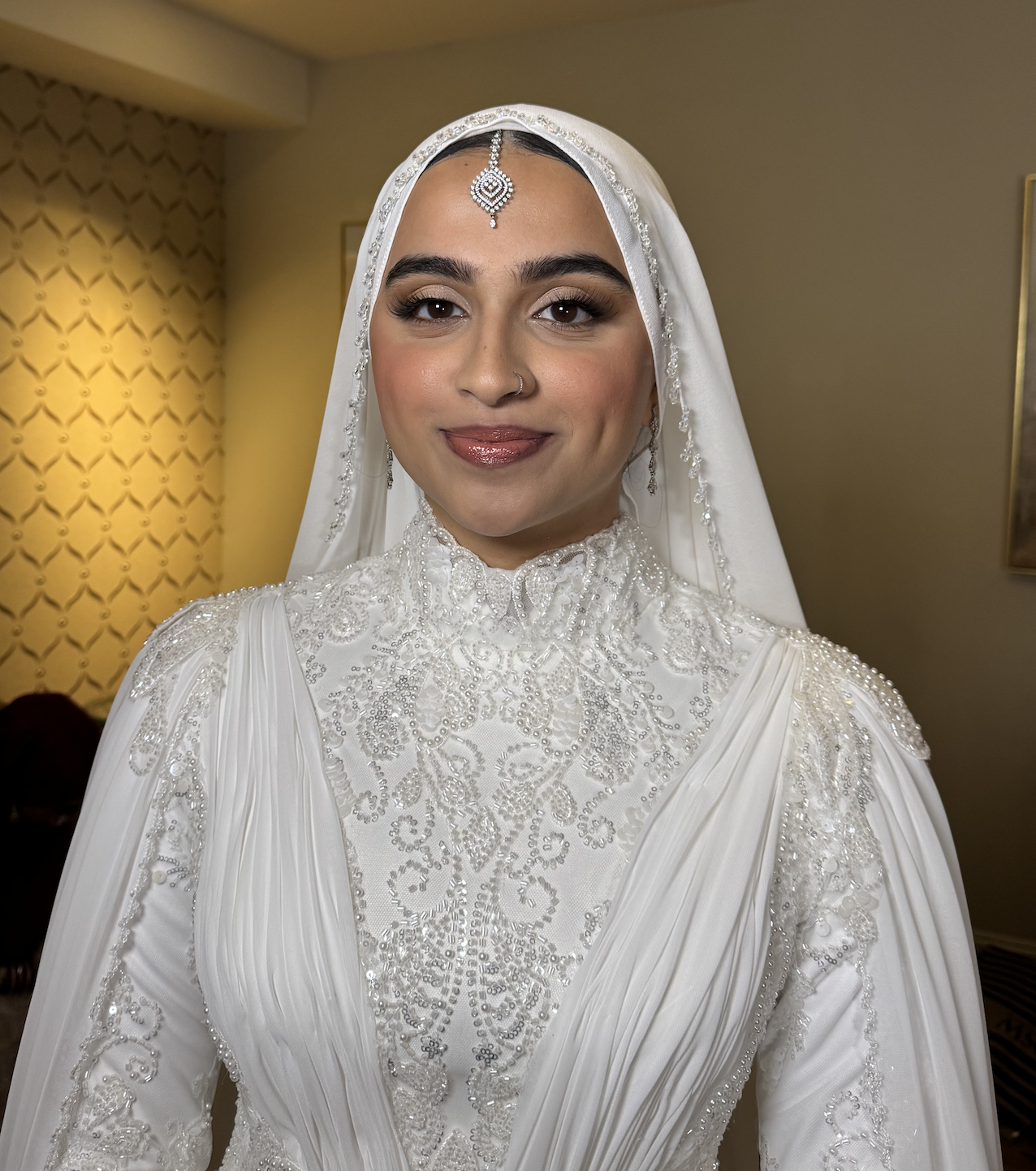 A woman in a white, ornate lace dress with pearl embellishments, wearing a matching veil and jewelry, smiling in an indoor setting.