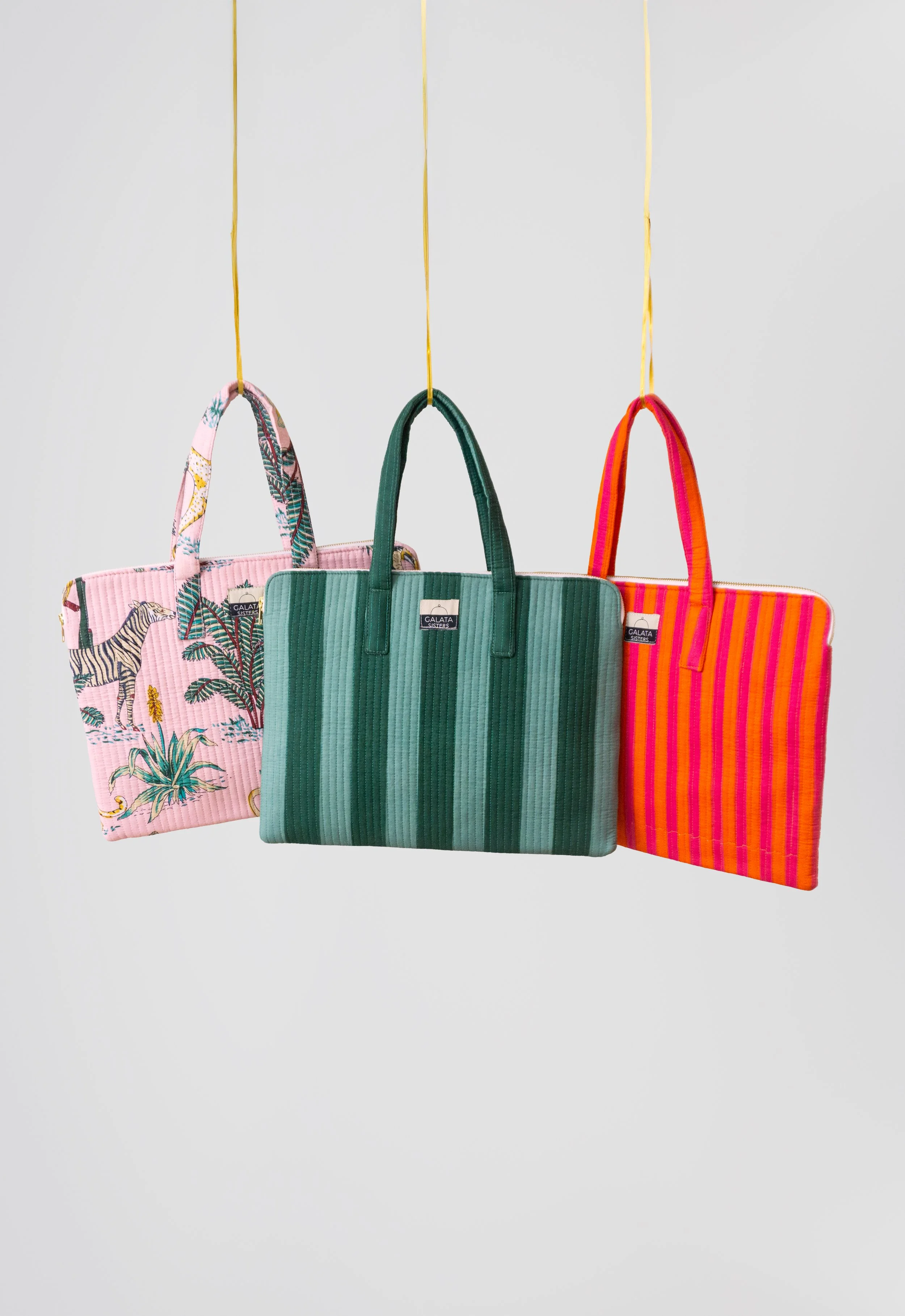 Tres bolsas de mano colgadas en cuerdas amarillas contra un fondo blanco, una con diseño de selva y animales, otra en tonos verdes y otra en rayas rosa y naranja.