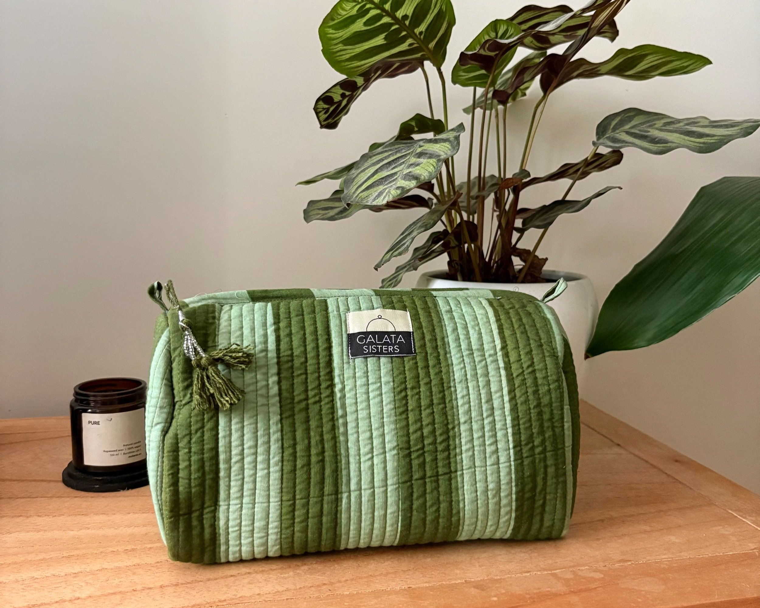 Una bolsa de tela verde con rayas en tonos de verde, con un lazo de cuerda y un etiqueta que dice 'Galata Sisters'. Está sobre una mesa de madera, junto a una vela negra y una planta en maceta blanca con hojas verdes y moradas.