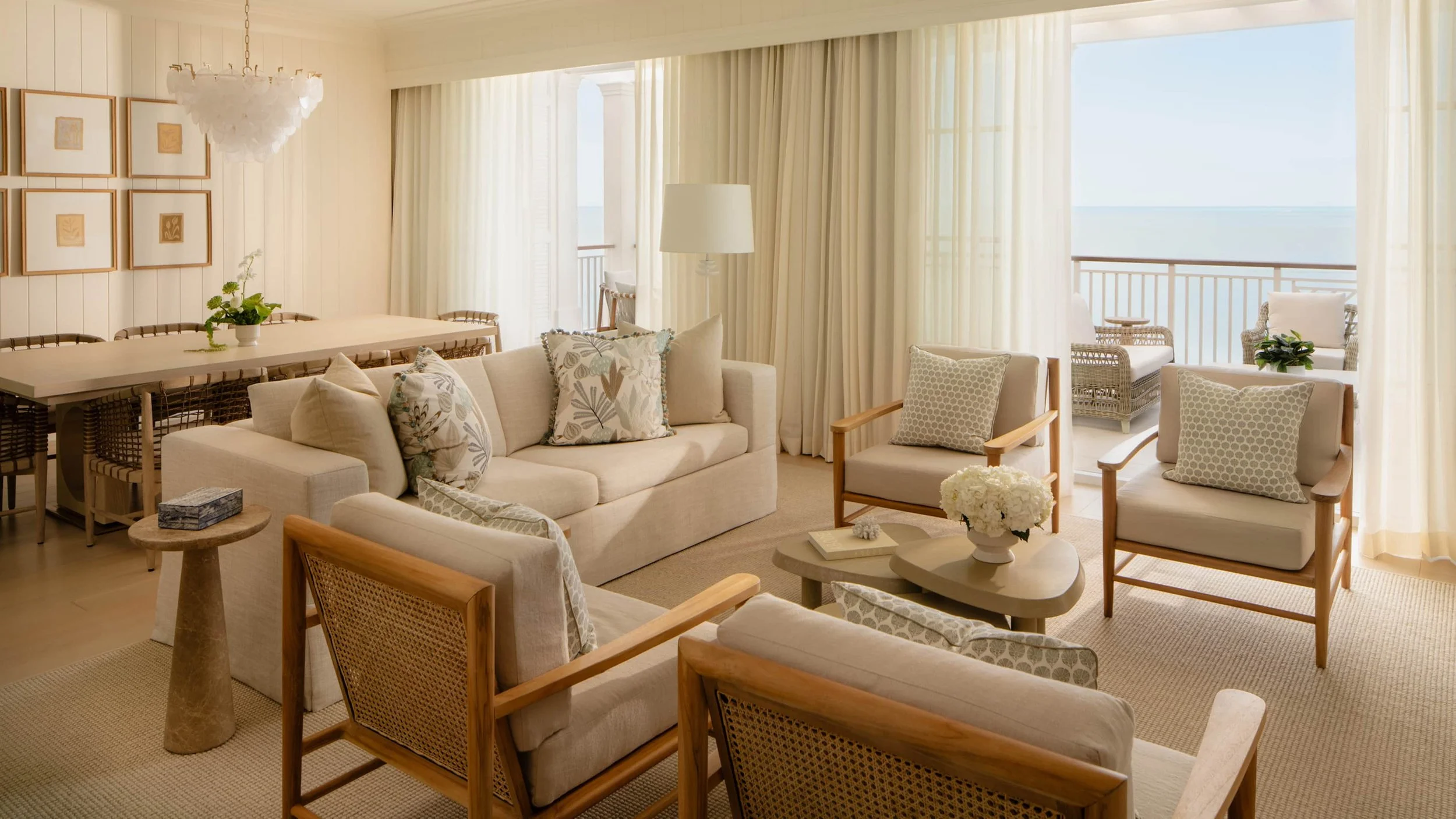 Living room with beige sofa set, wooden chairs, and glass doors opening to a balcony with ocean view, decorated with framed artwork, plants, and soft lighting.