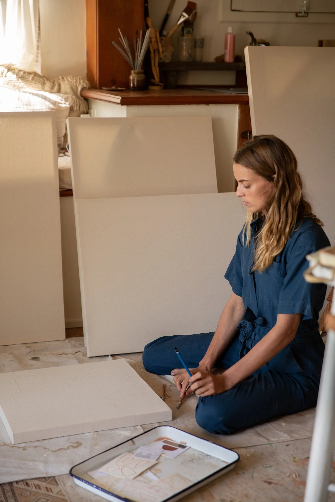 A woman sitting cross-legged on the floor, drawing on a large sheet of paper with a pencil. There are several foam boards or canvases behind her, and art supplies are visible in the background on a shelf. A tray with sketches or notes is on the floor nearby.