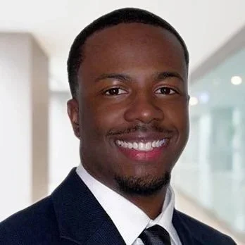 A young man with dark skin, wearing a navy blue suit and a black tie, smiling in a modern office setting with glass walls and a hallway in the background.