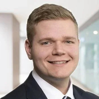 A young man with blonde hair wearing a black suit, white shirt, and striped tie, smiling in a modern office with glass walls and a wooden wall in the background.