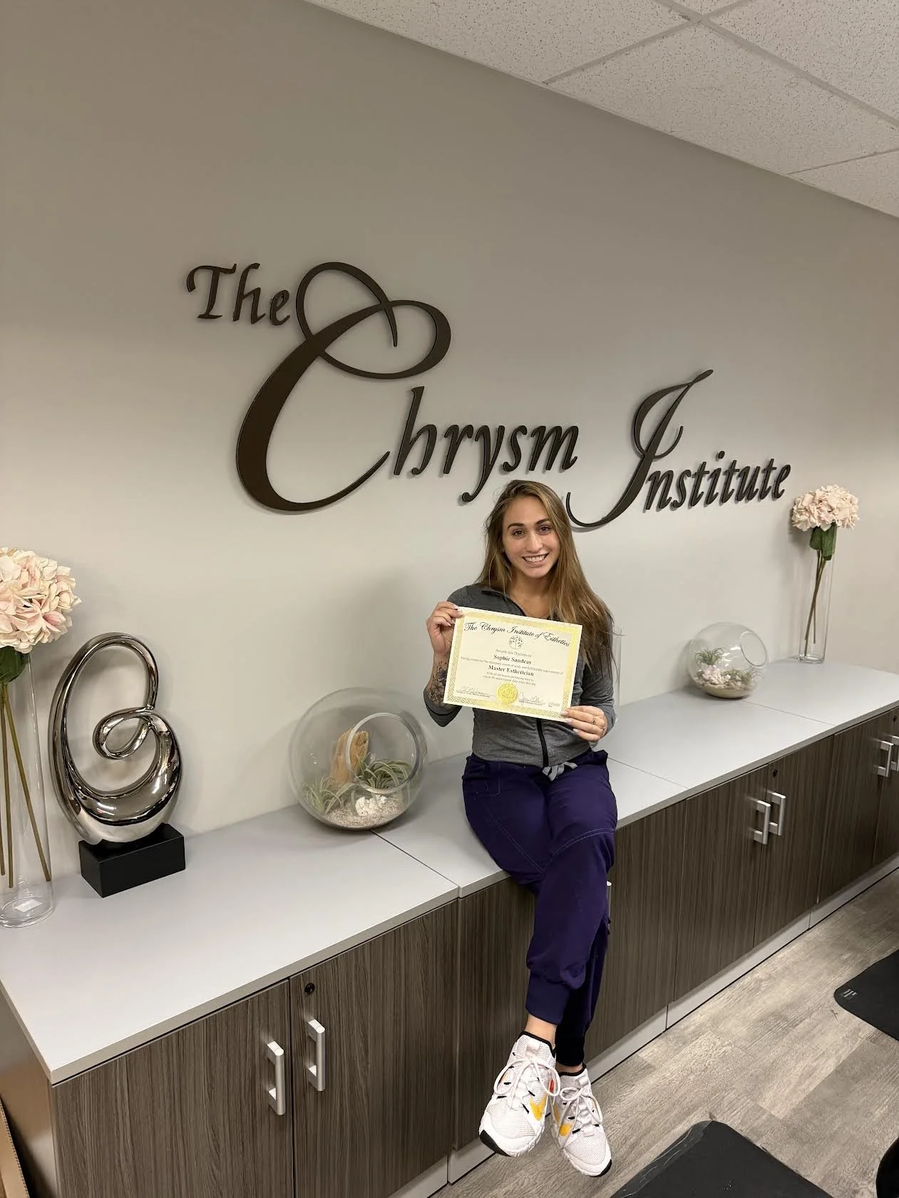 Young woman sitting on a counter, smiling and holding a certificate in front of a wall with the sign 'The Chrysma Institute' and decorative flowers.