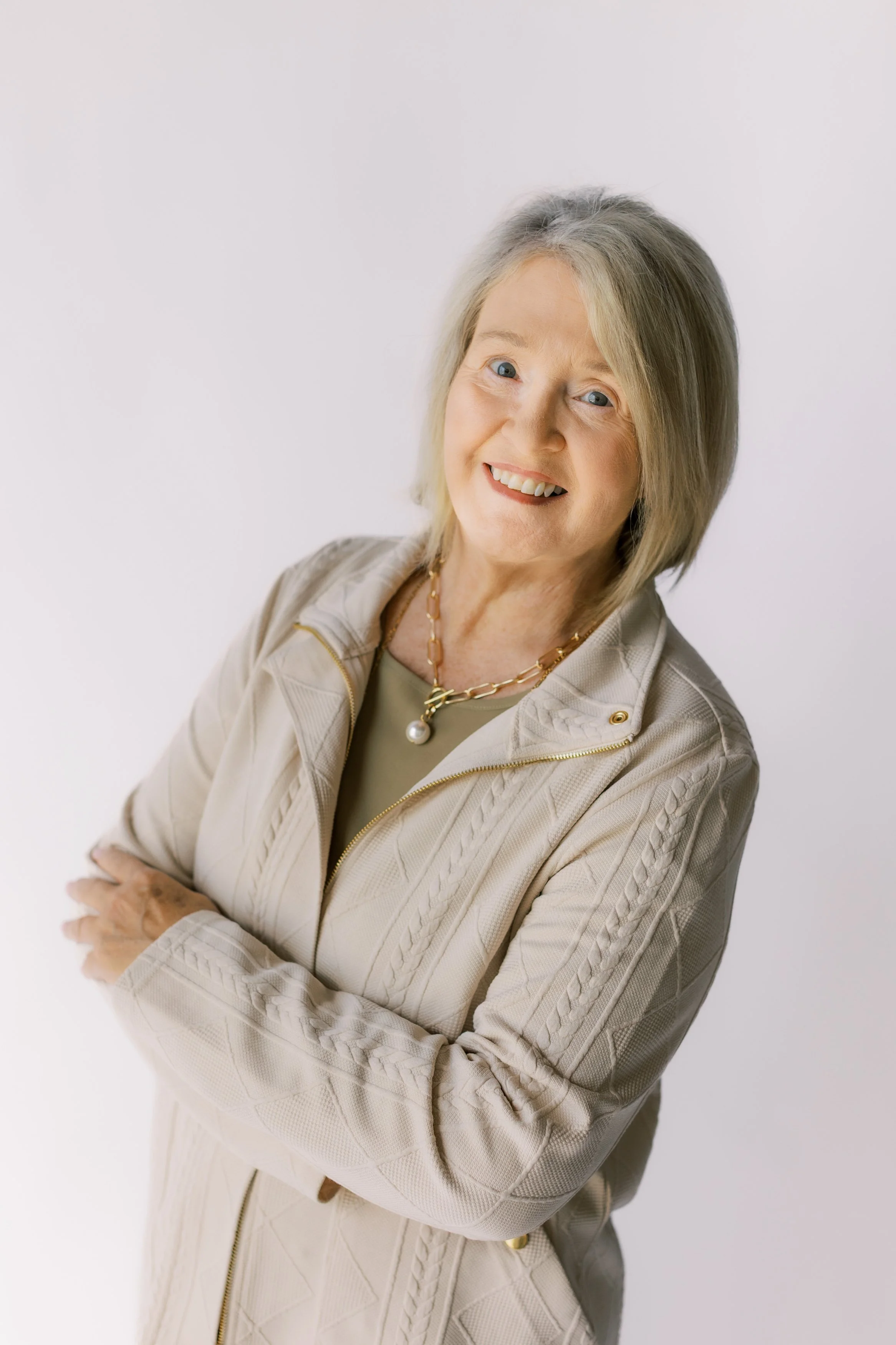 An older woman with short gray hair, wearing a black blazer, smiling and standing against a plain light-colored wall.