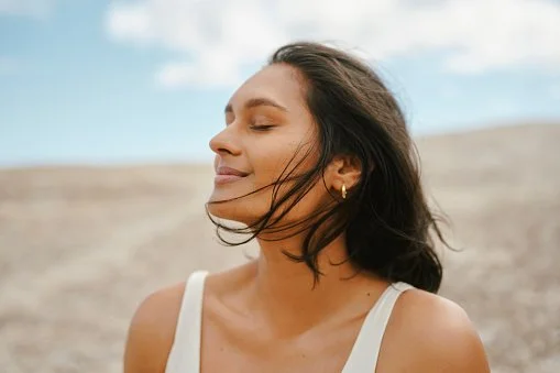 A woman with dark hair and earrings is outdoors with a desolate landscape and a partly cloudy sky in the background.
