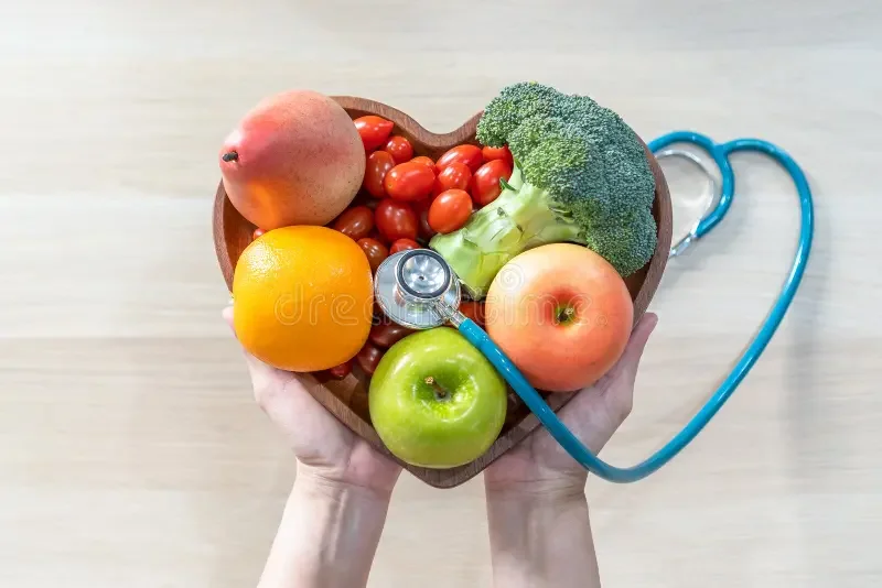 Hands holding a heart-shaped wooden bowl filled with various fruits and vegetables, including an apple, tomato, broccoli, orange, peach, cherry tomatoes, and a stethoscope resting on the bowl.