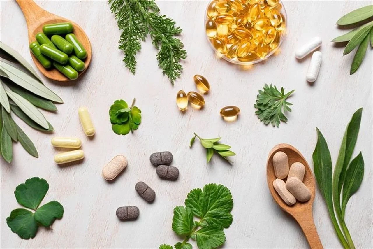 Assorted herbal supplements and capsules spread on a light wooden surface with green leaves and sprigs of herbs.