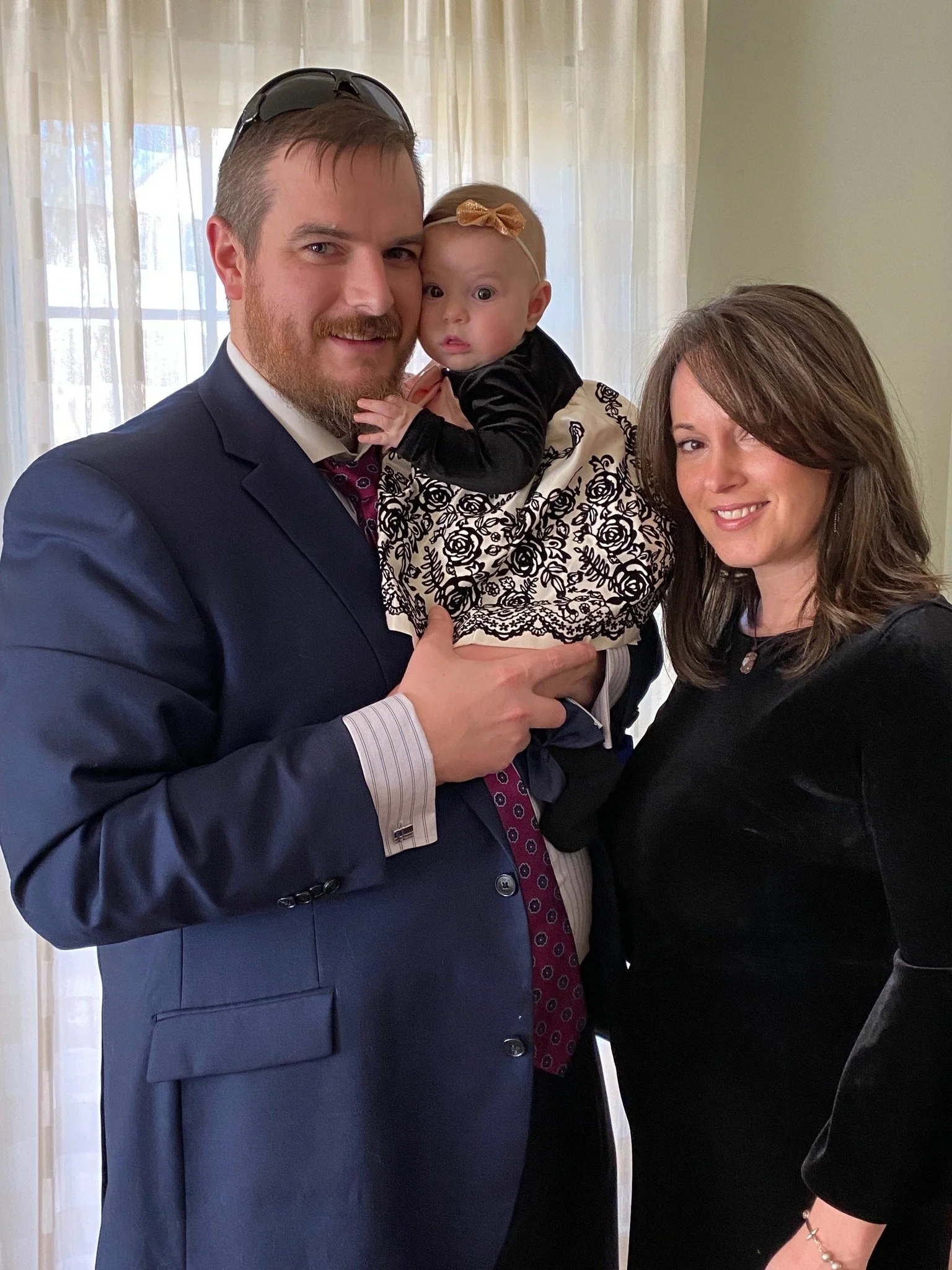 A man in a navy suit holding a baby girl, with a woman standing beside them. The baby is wearing a cream dress with black floral lace overlay and a gold bow headband. The man has sunglasses on his head, and they are indoors near a window with sheer curtains.