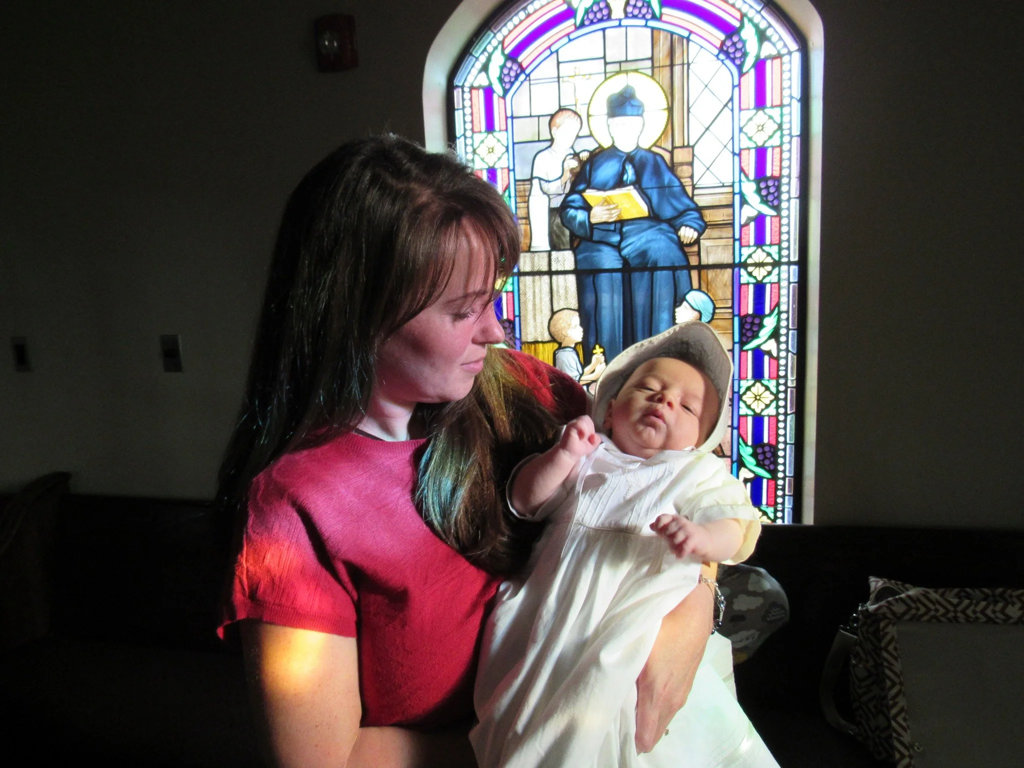 A woman with long hair holding a smiling baby in front of a colorful stained-glass window in a church.