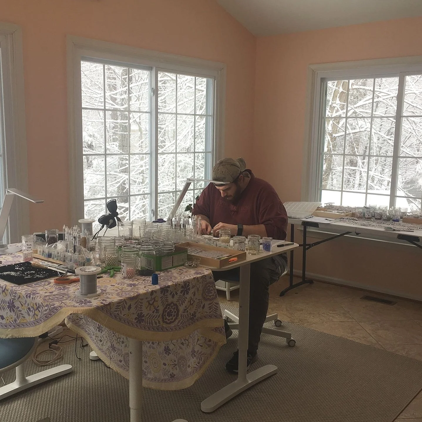 A person seated at a table, working with small items in a room with large windows showing a snowy landscape outside. The table is covered with jars, tools, and supplies.