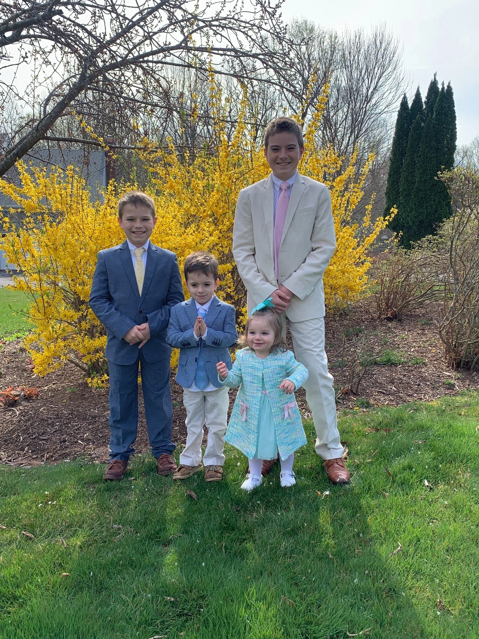Four children dressed in formal clothing standing outdoors in front of a yellow flowering shrub, with leafless trees and tall green shrubs in the background.