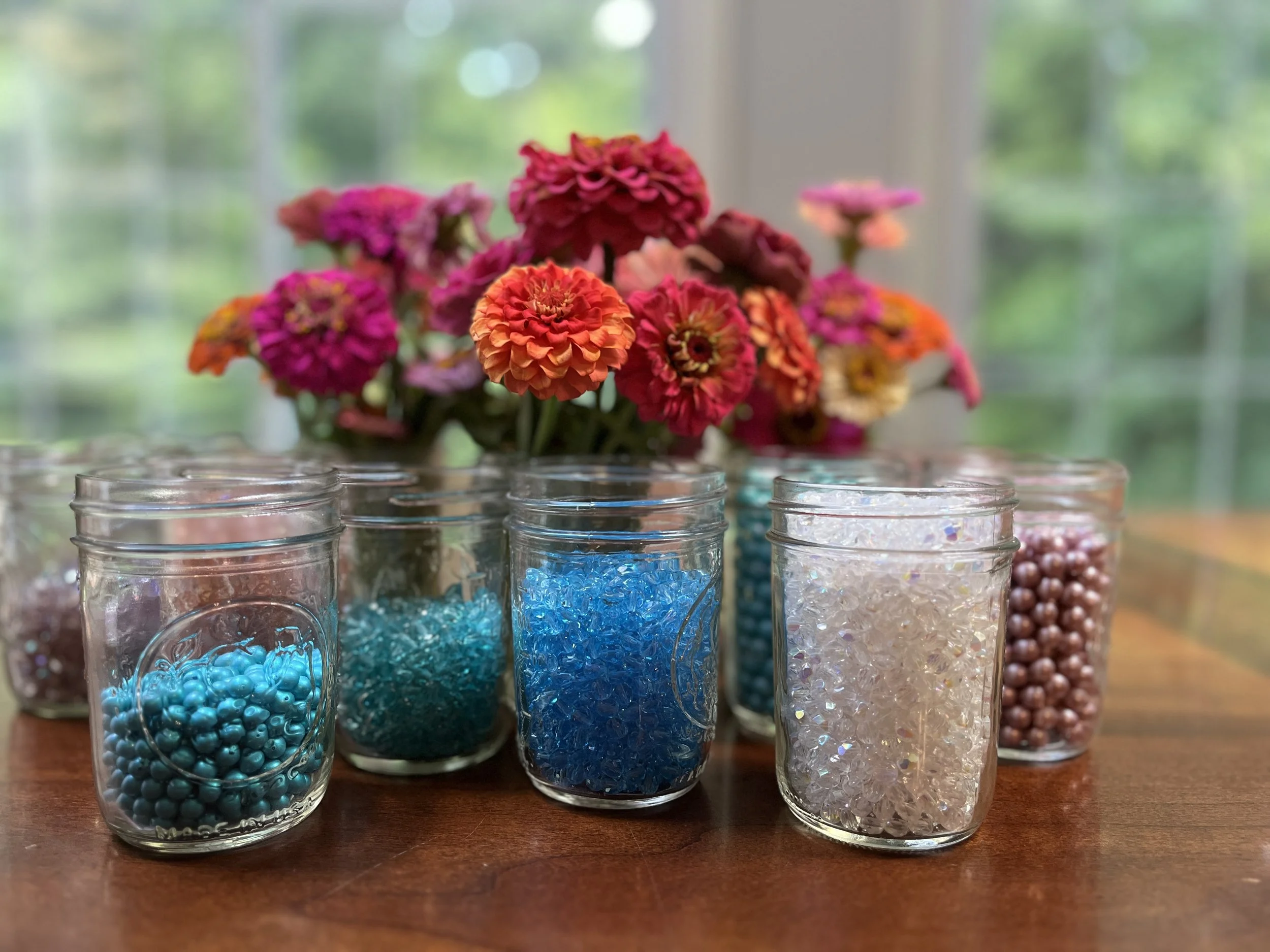 A bouquet of pink, orange, and red flowers on a wooden table with glass jars filled with colorful beads and small decorative items in the foreground, and a blurred background of a window with greenery outside.