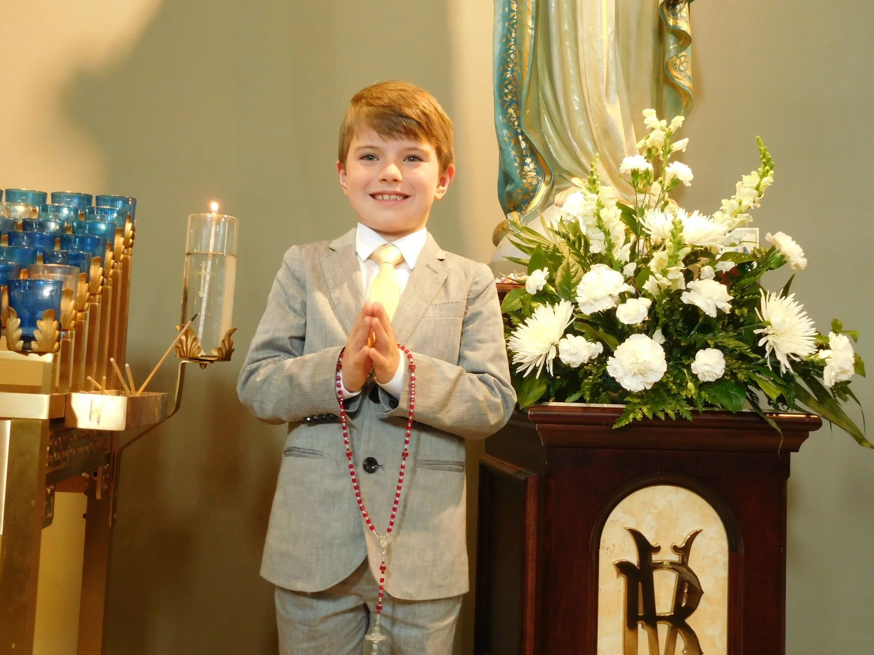 Young boy dressed in a gray suit, standing with hands pressed together in prayer, smiling, in front of a religious altar with white flowers and a statue.