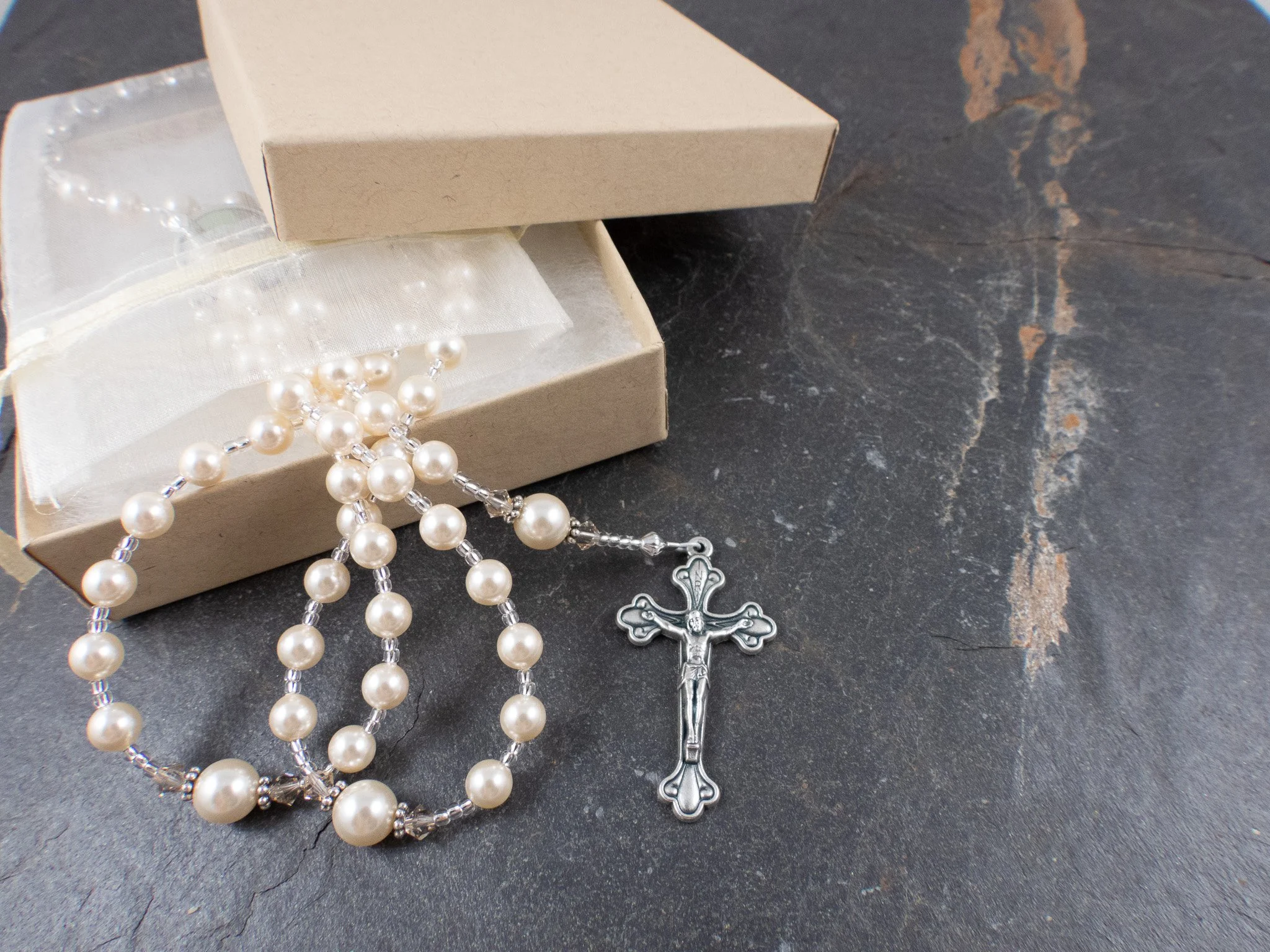 A jewelry box with a string of pearl and crystal beads, a silver cross crucifix, and a white fabric pouch on a dark textured surface.