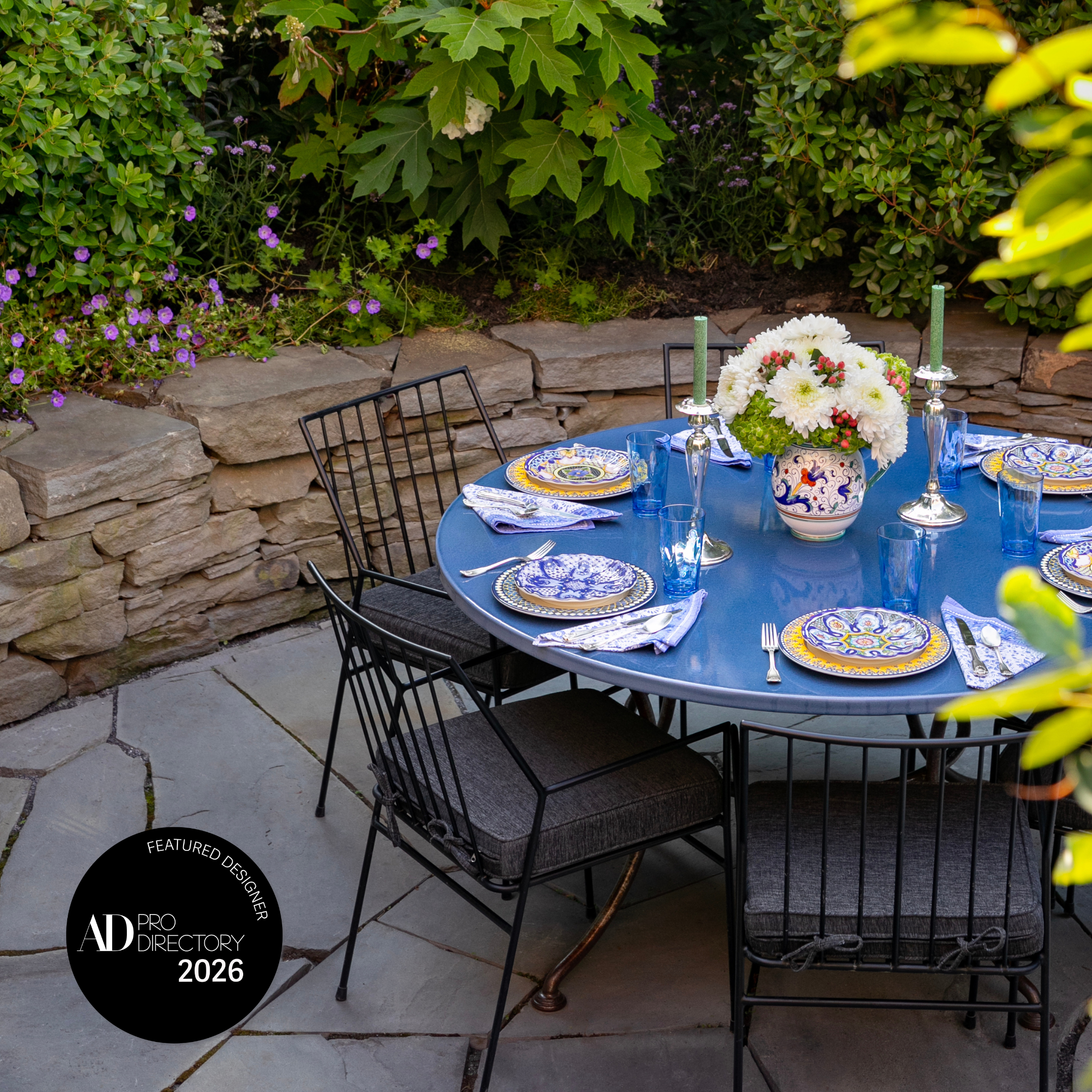 Fort Greene NYC Brooklyn Brownstone backyard featuring a stacked stone wall, hand laid bluestone pavers, and natural upstate woodland trees and plants. An elegant metal patio set and blue enamel table are set. AD Pro directory 2026.