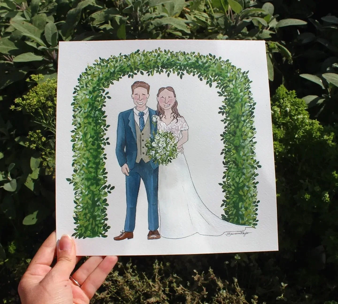 Wedding invitation and detail card with floral wreath and the names 'Ewan and Ffion' written in calligraphy, placed on grass and surrounded by green plants.