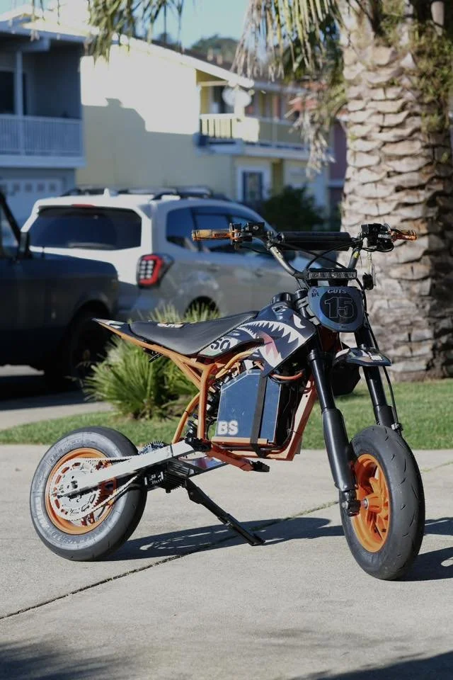 Electric dirt bike with Shark on it, parked on a sidewalk in a residential neighborhood with palm trees and houses in the background.