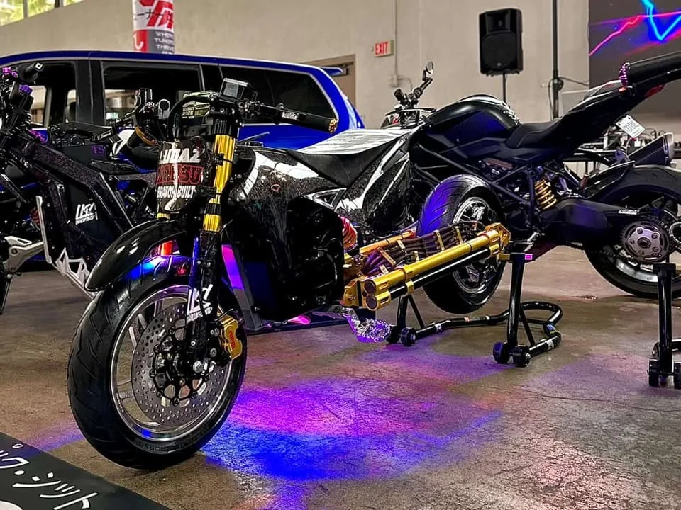 A futuristic motorcycle with black and gold components displayed indoors, with other motorcycles and a vehicle visible in the background.