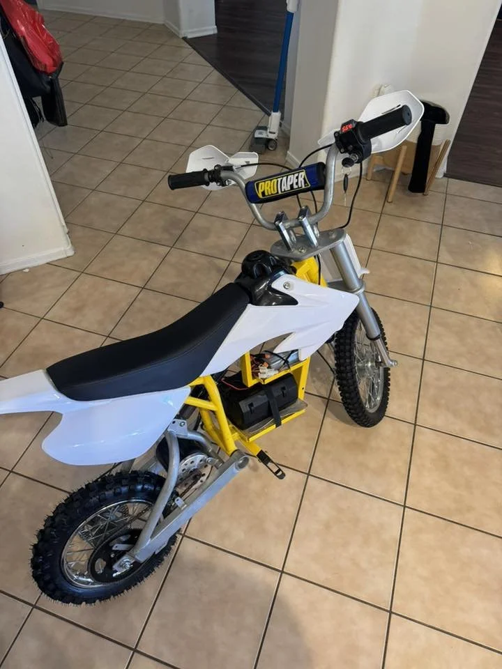 A small dirt bike with a yellow frame, black seat, and white fenders, positioned on a tiled floor indoors.