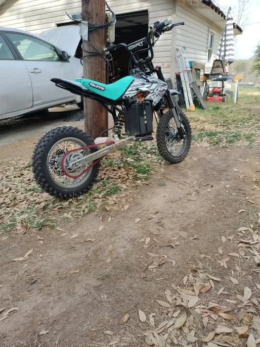 A small dirt bike with a teal seat and black frame leaning against a wooden utility pole in a yard with a car, a house, and outdoor furniture in the background.