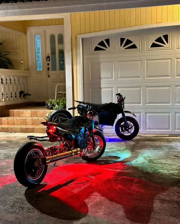 Two motorcycles parked in a garage with colored lights reflecting on the concrete floor.