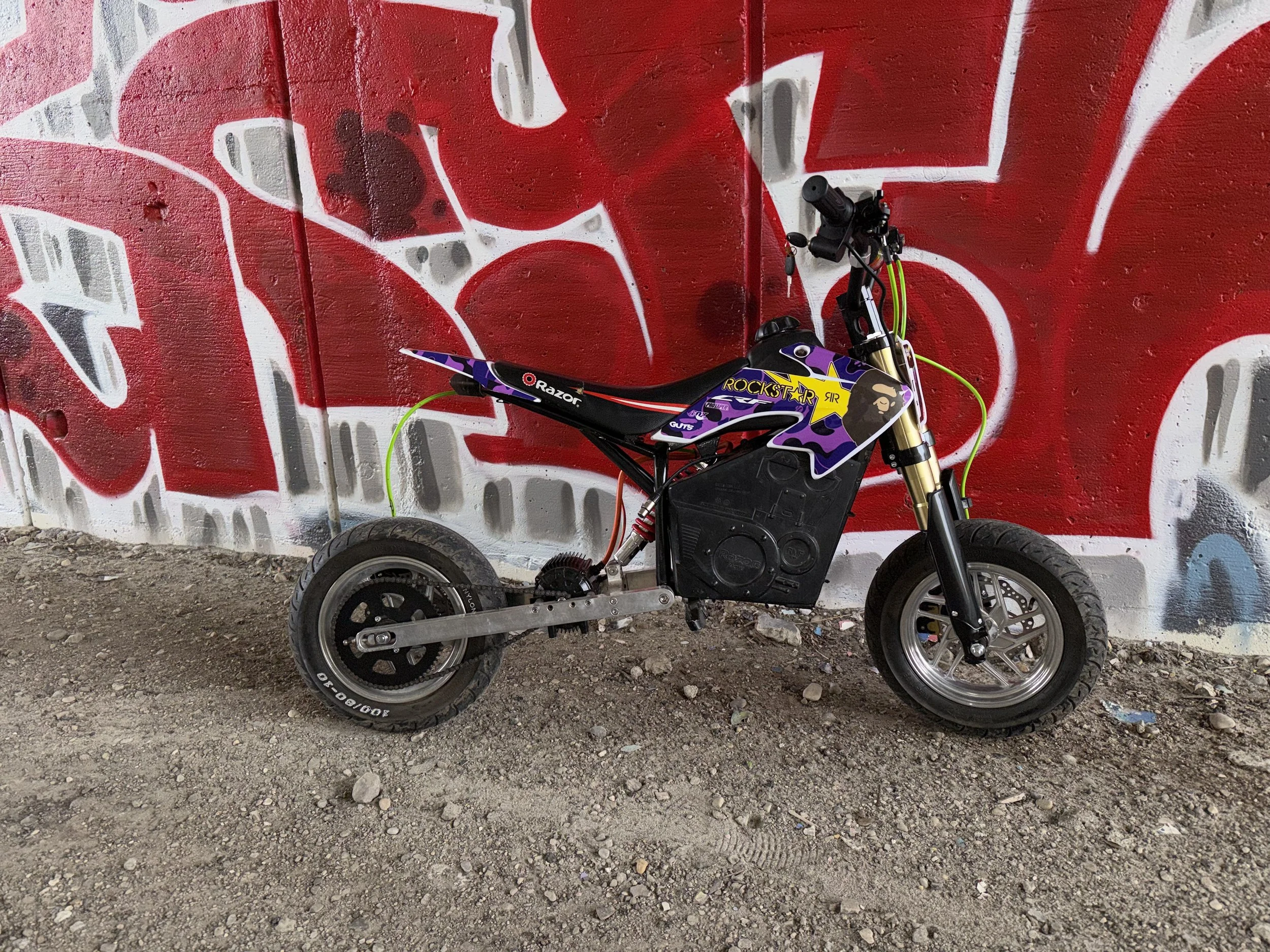 A small electric motorcycle with a black body, purple and yellow graphics, and a 'Rockstar' logo, parked on gravel in front of a graffiti-covered wall.