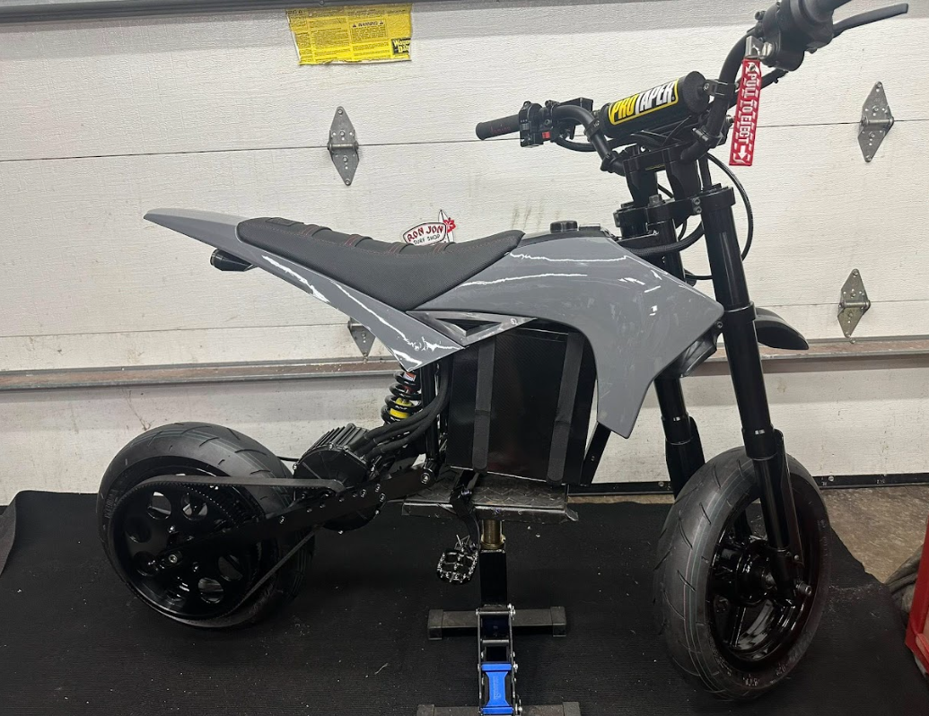 A gray electric motorcycle with a black seat, large tires, and a visible suspension system, positioned in a garage with a white garage door in the background.