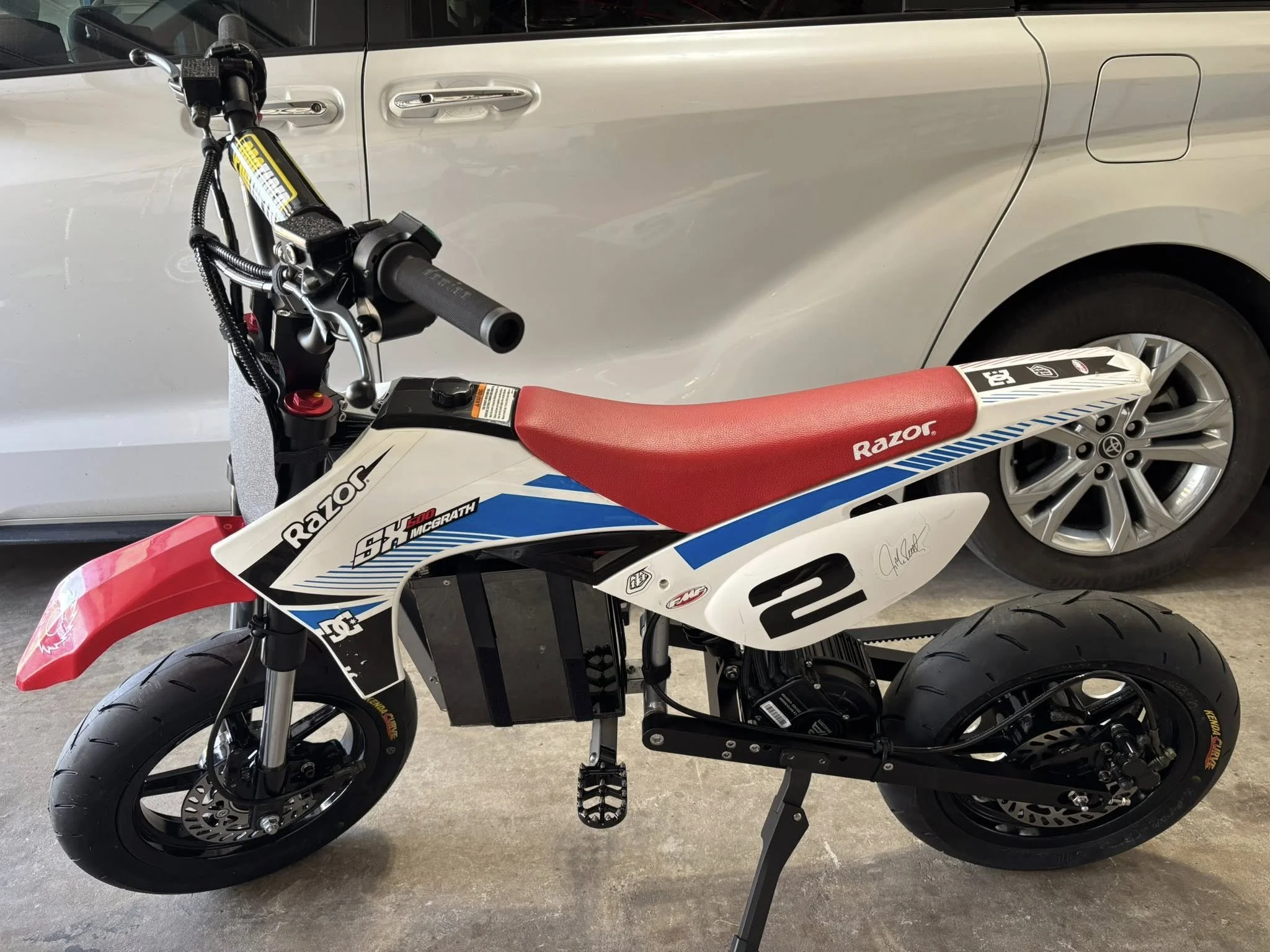 A small electric dirt bike with a red seat, black frame, and racing decals, parked in a garage with a silver car in the background.